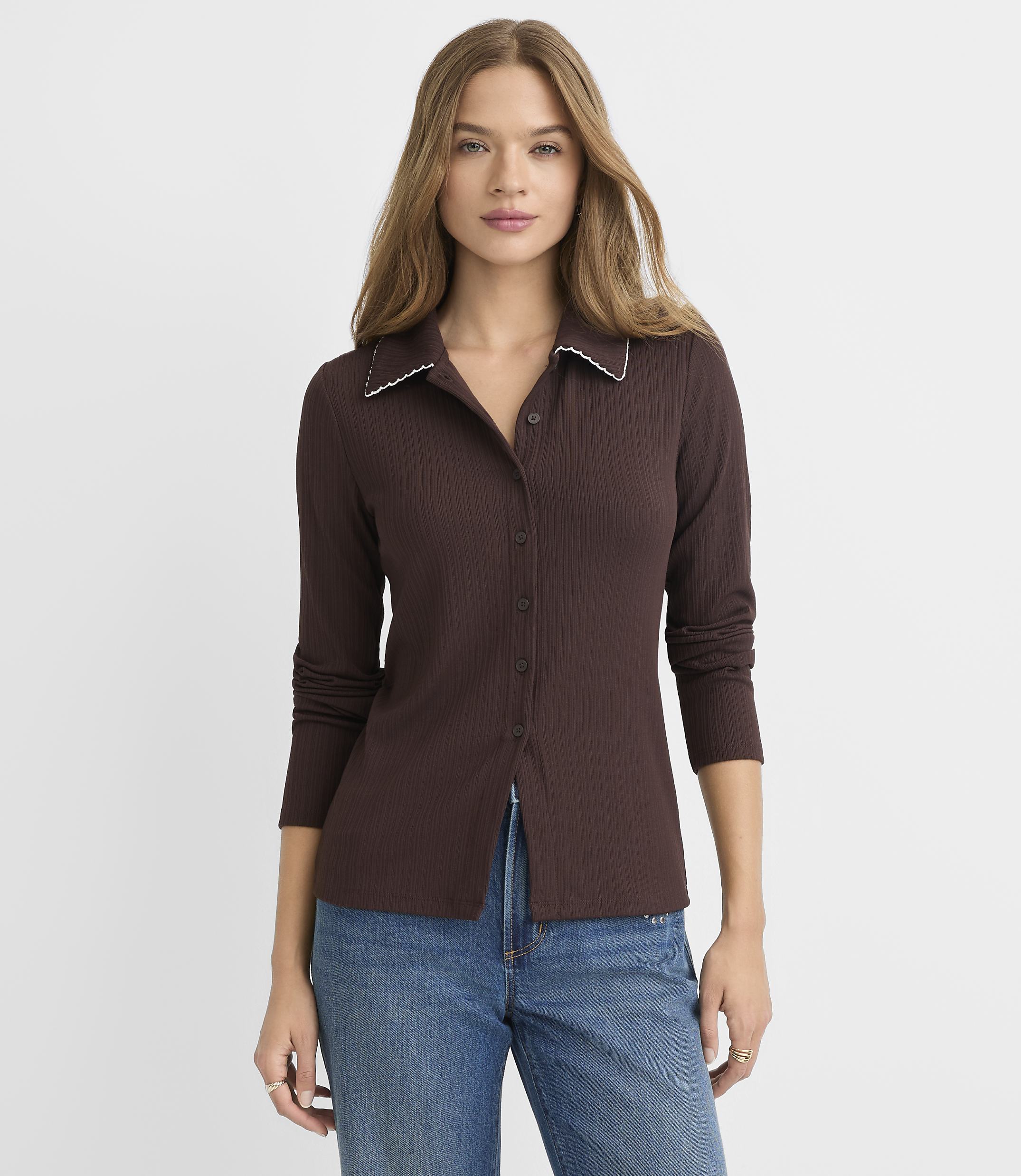 A woman in a brown ribbed collared shirt and blue jeans stands against a plain white background.