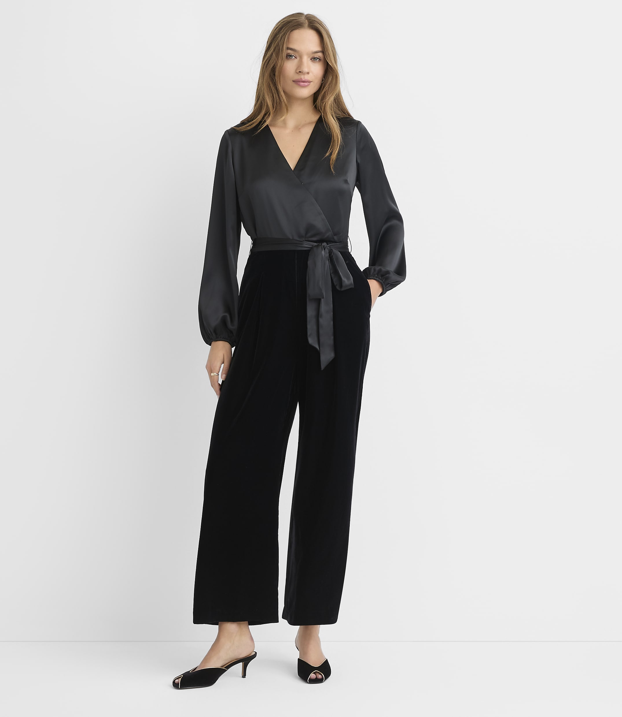 A woman in a black satin wrap top and velvet pants stands against a plain white background.