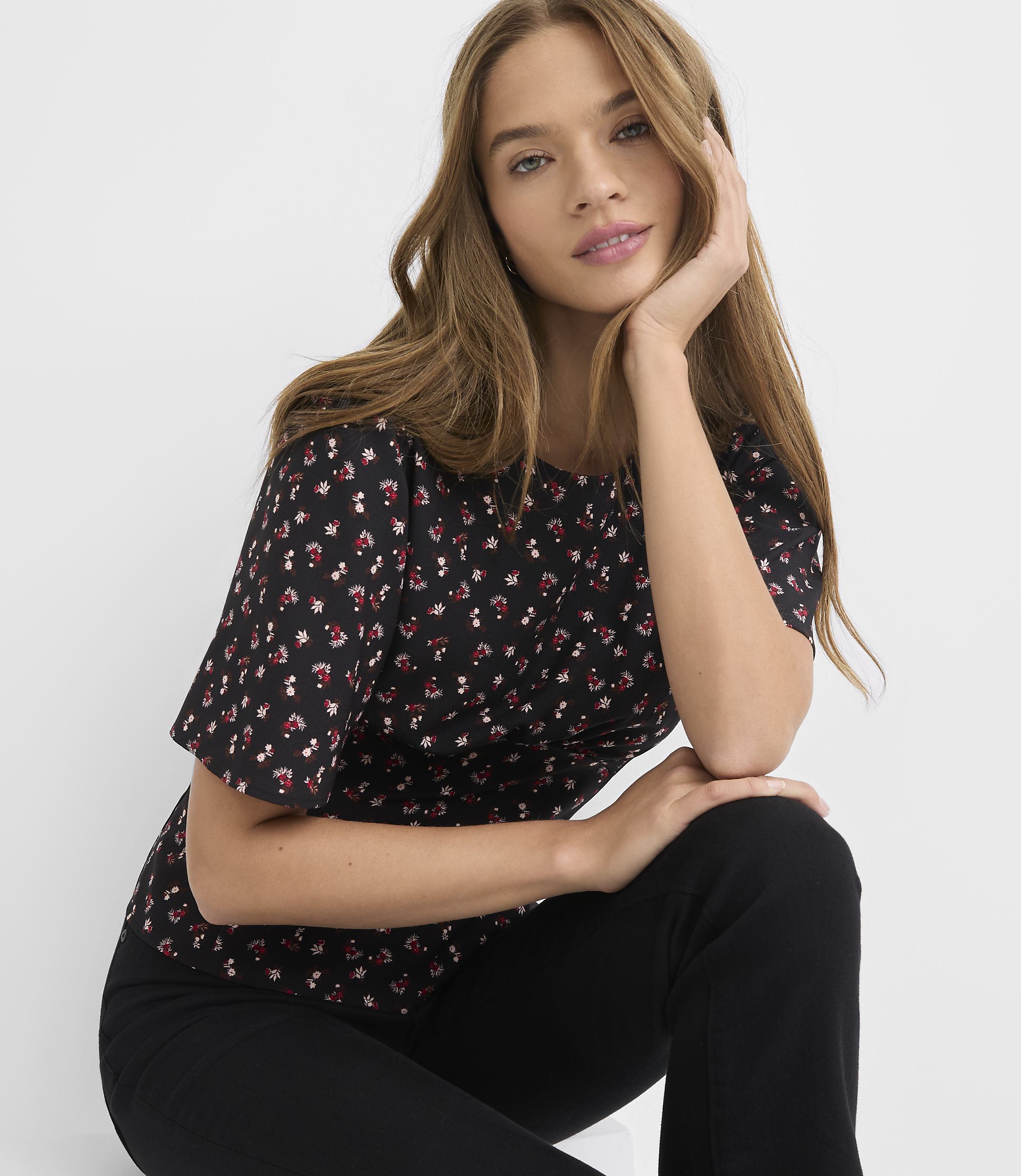 A young woman with long brown hair, wearing a black floral t-shirt and black pants, poses against a plain white background.
