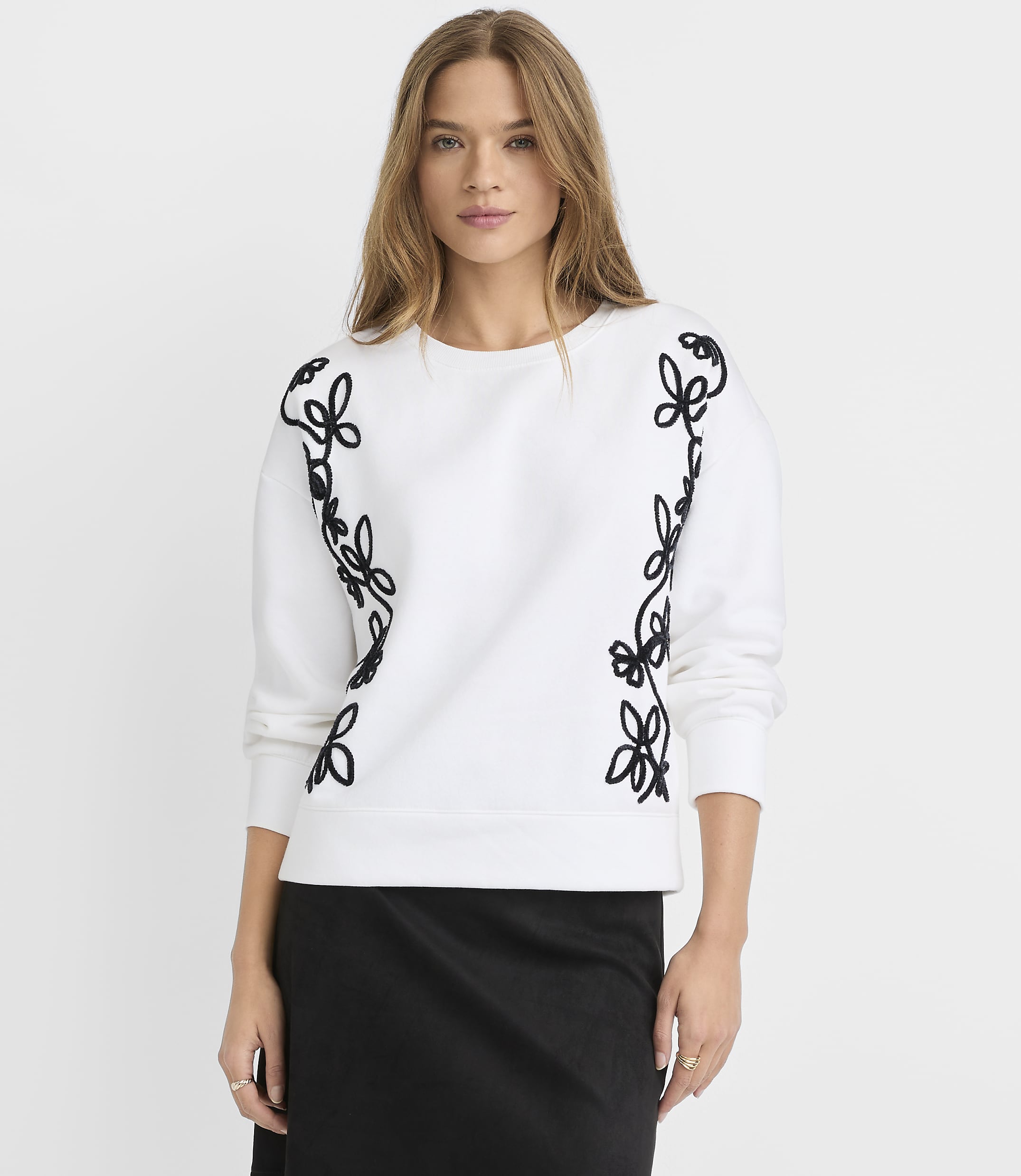 A woman in a white sweatshirt with black floral embroidery stands against a plain white background.