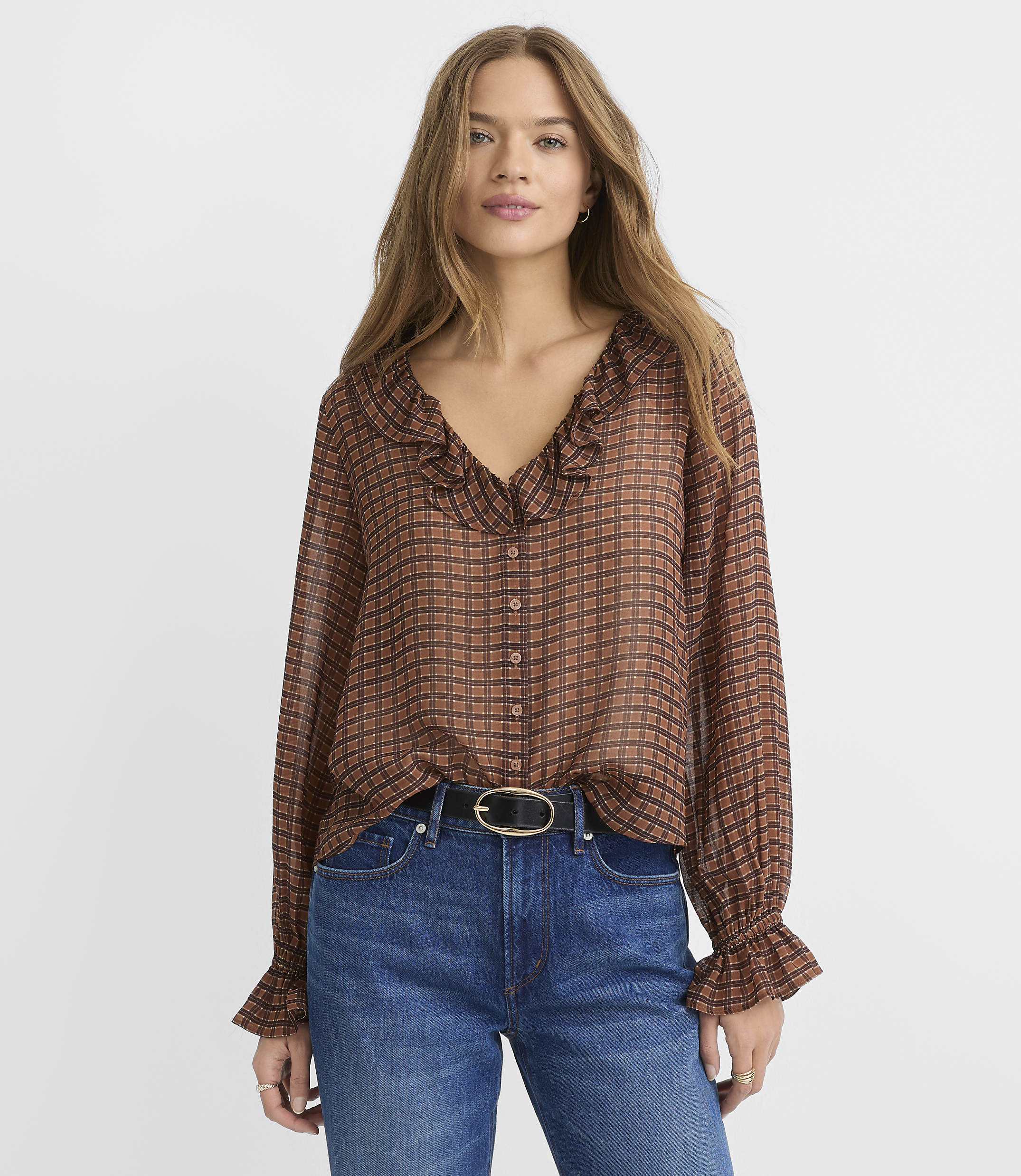 A woman in a brown checkered blouse and blue jeans stands against a plain white background.