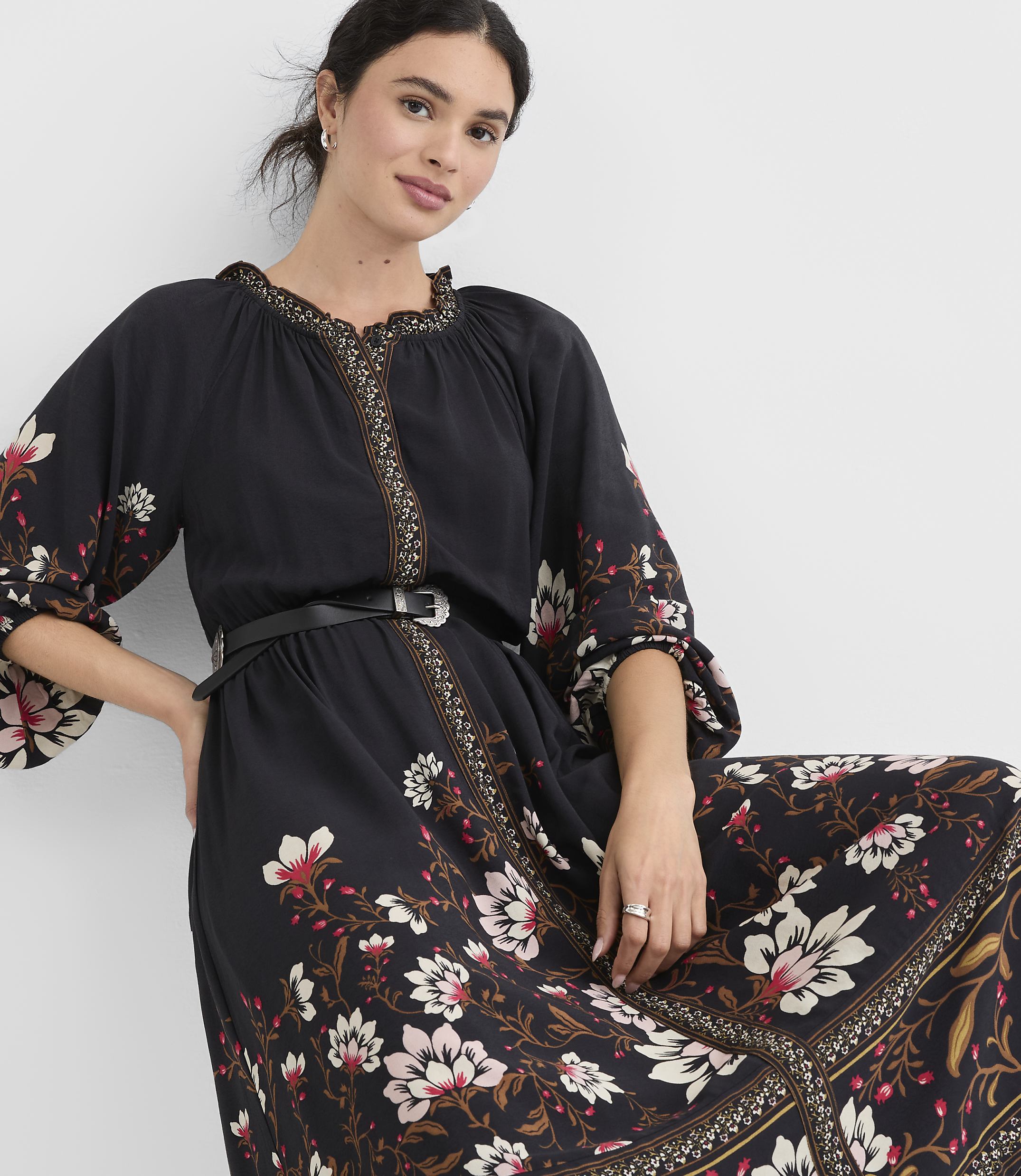 A woman in a black floral dress with a cinched waist stands against a plain white background.