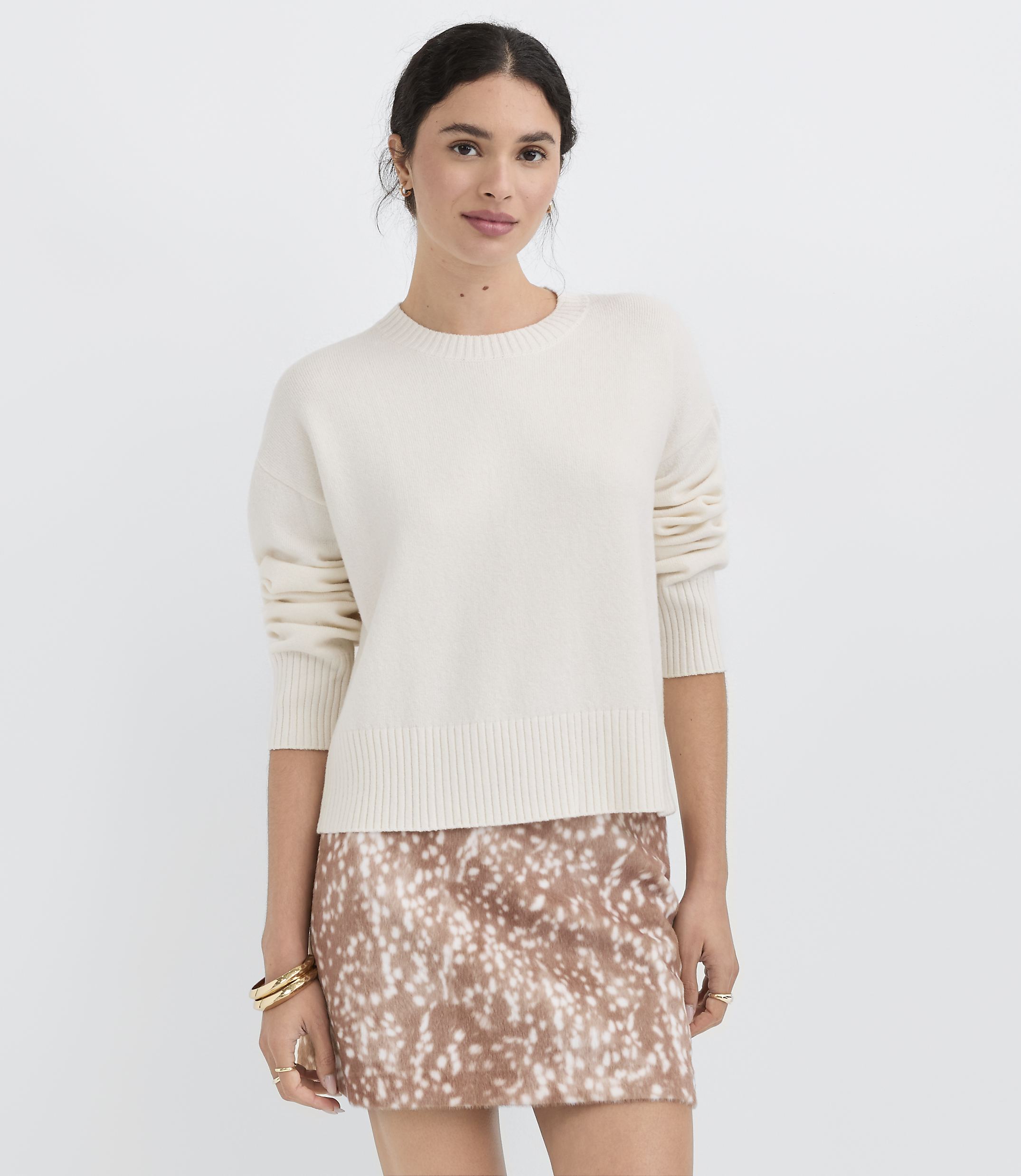 A woman in a cream sweater and a patterned skirt stands against a plain white background.
