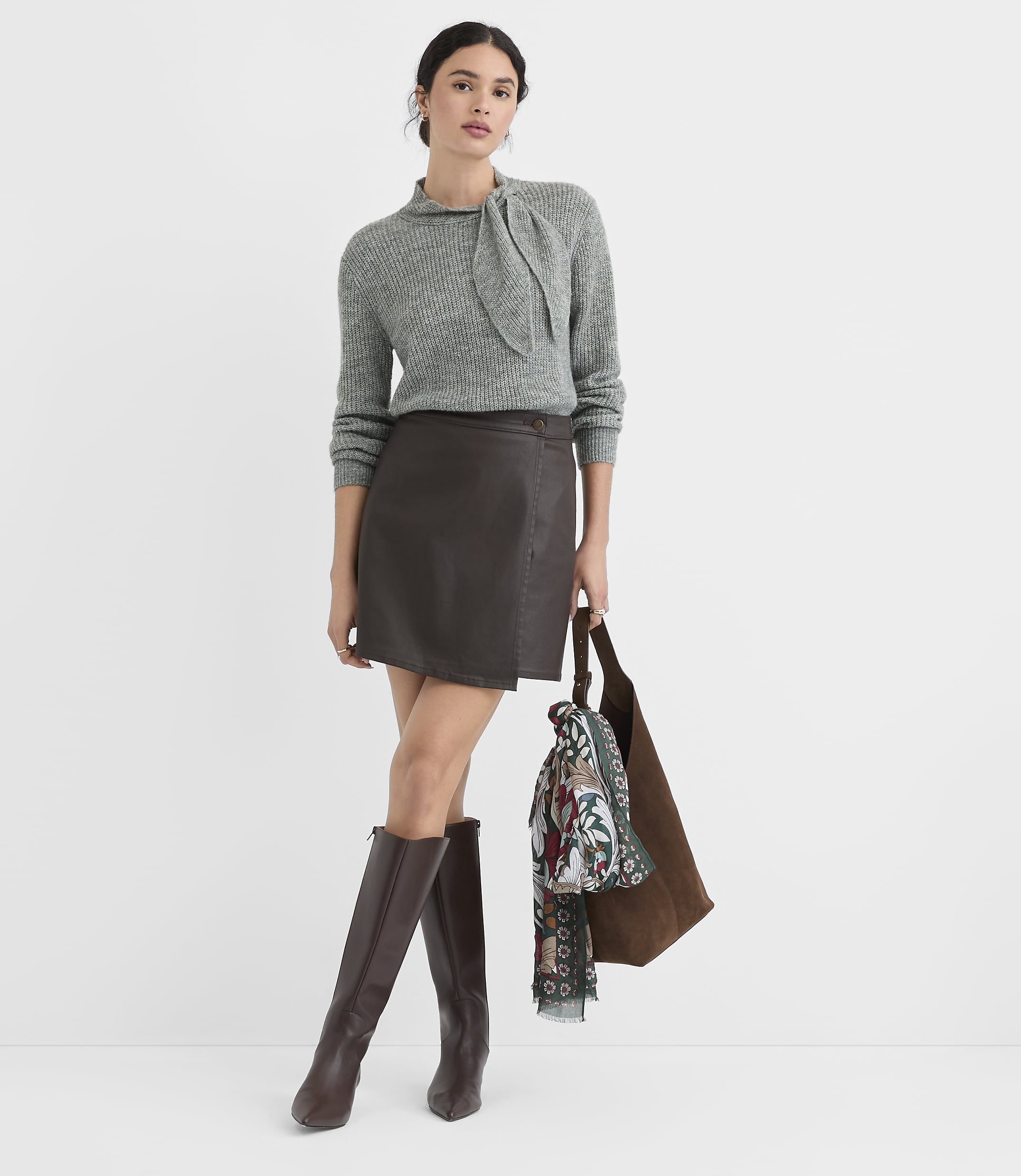 A woman in a grey sweater, brown leather skirt, and knee-high boots stands against a plain white background, holding a brown tote bag with a scarf draped over it.