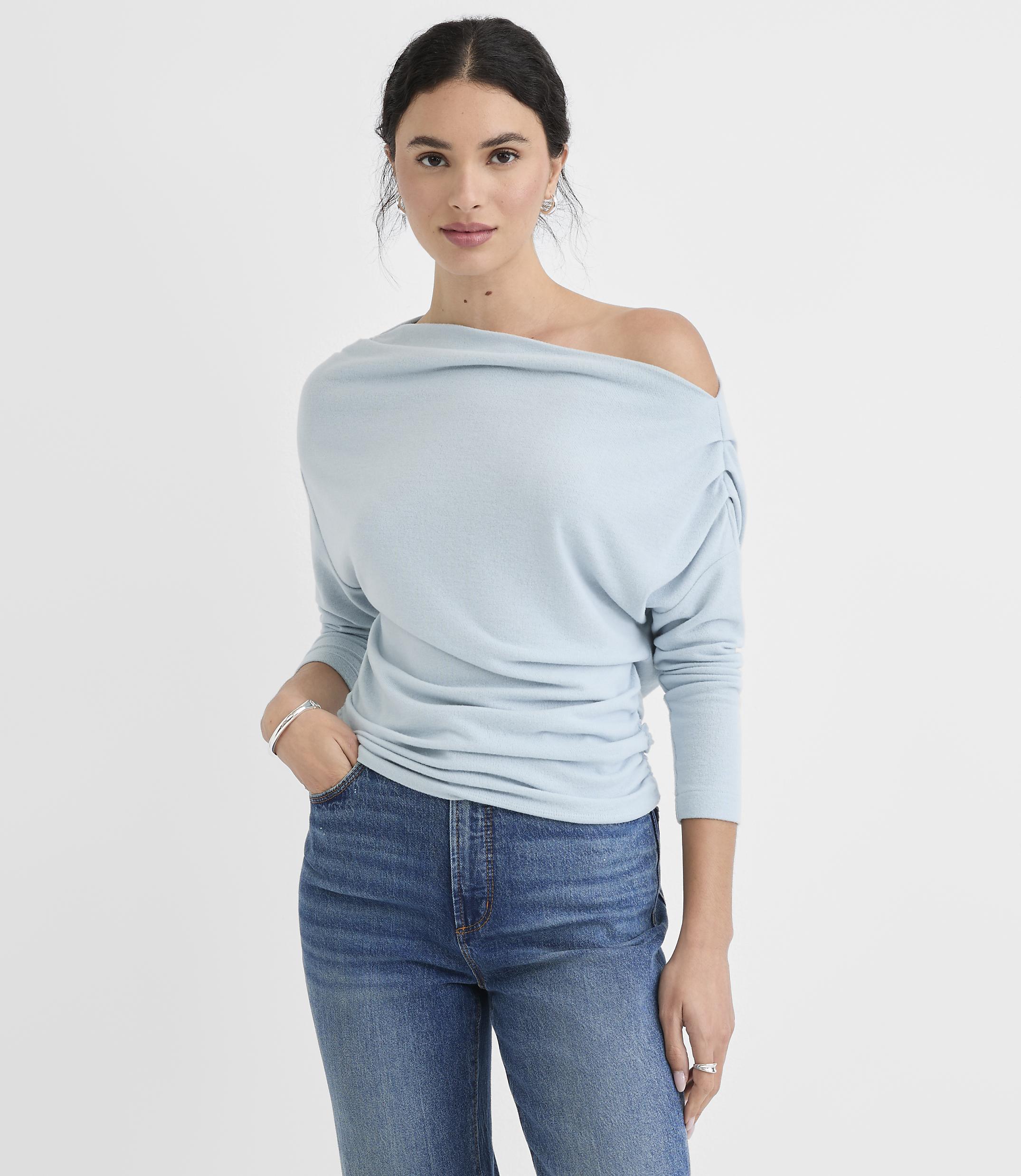 A woman in a light blue off-the-shoulder top and jeans stands against a plain white background.