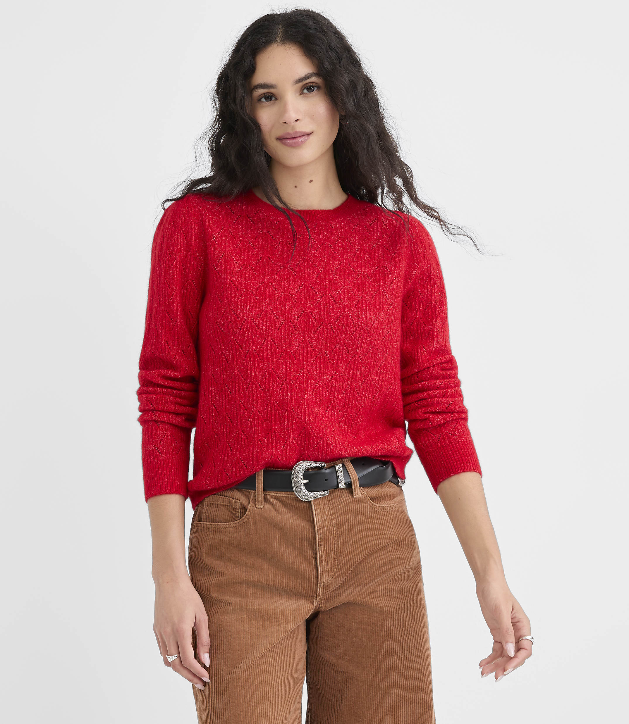 A woman in a red sweater and brown pants stands against a plain white background.