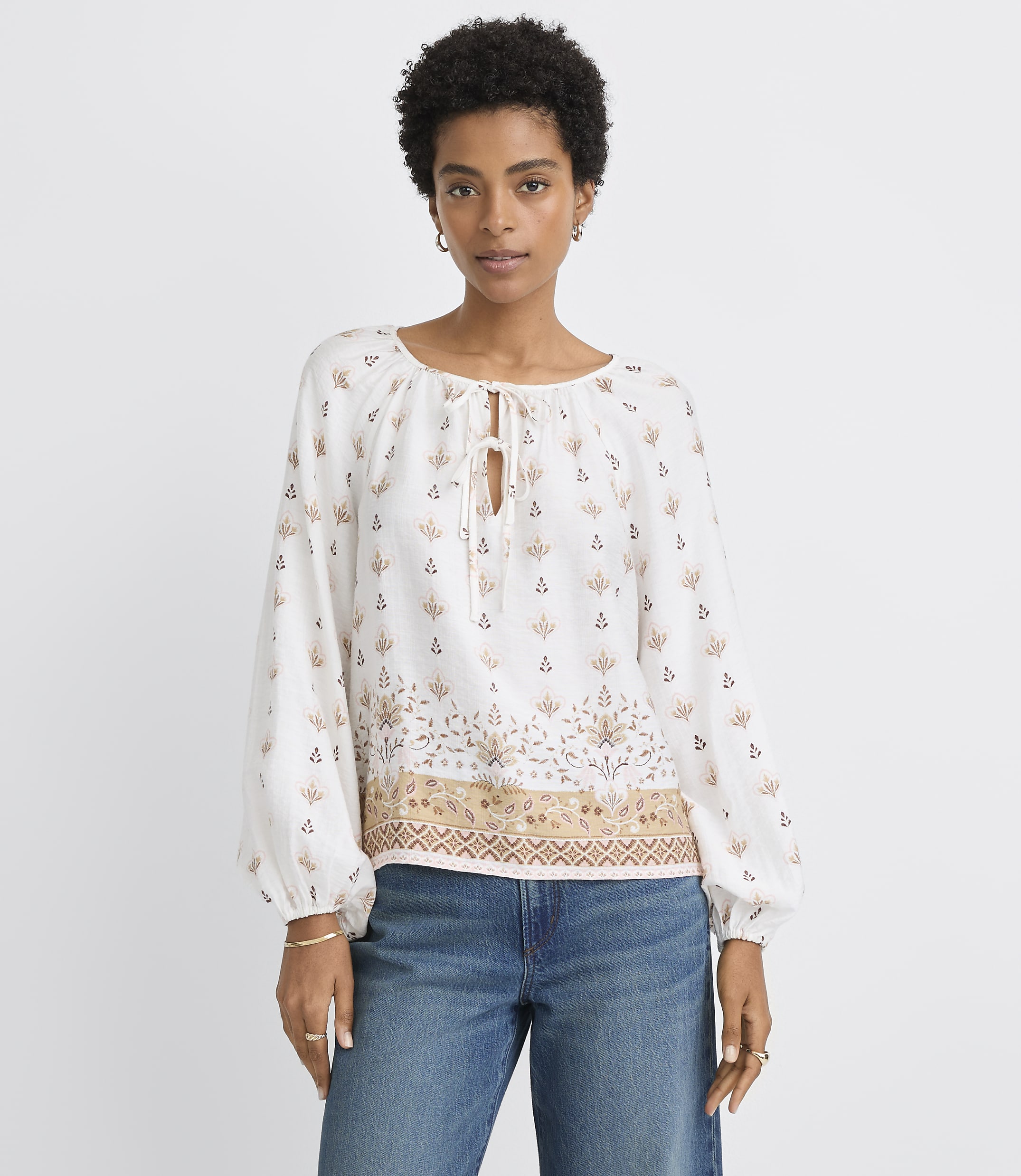 A woman in a patterned white blouse and blue jeans stands against a plain white background.
