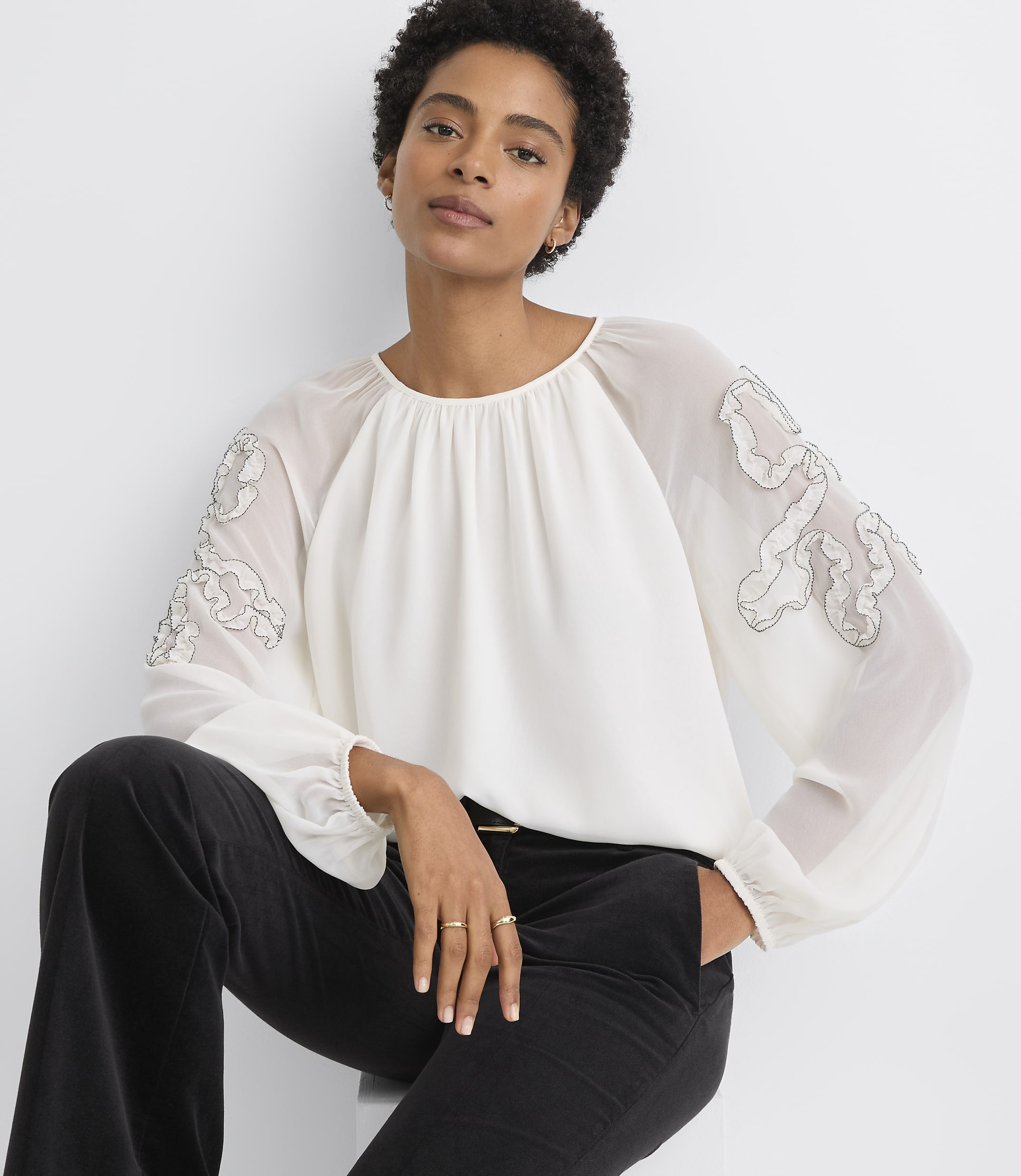 A woman in a white blouse with sheer, embroidered sleeves and black pants sits against a plain white background.