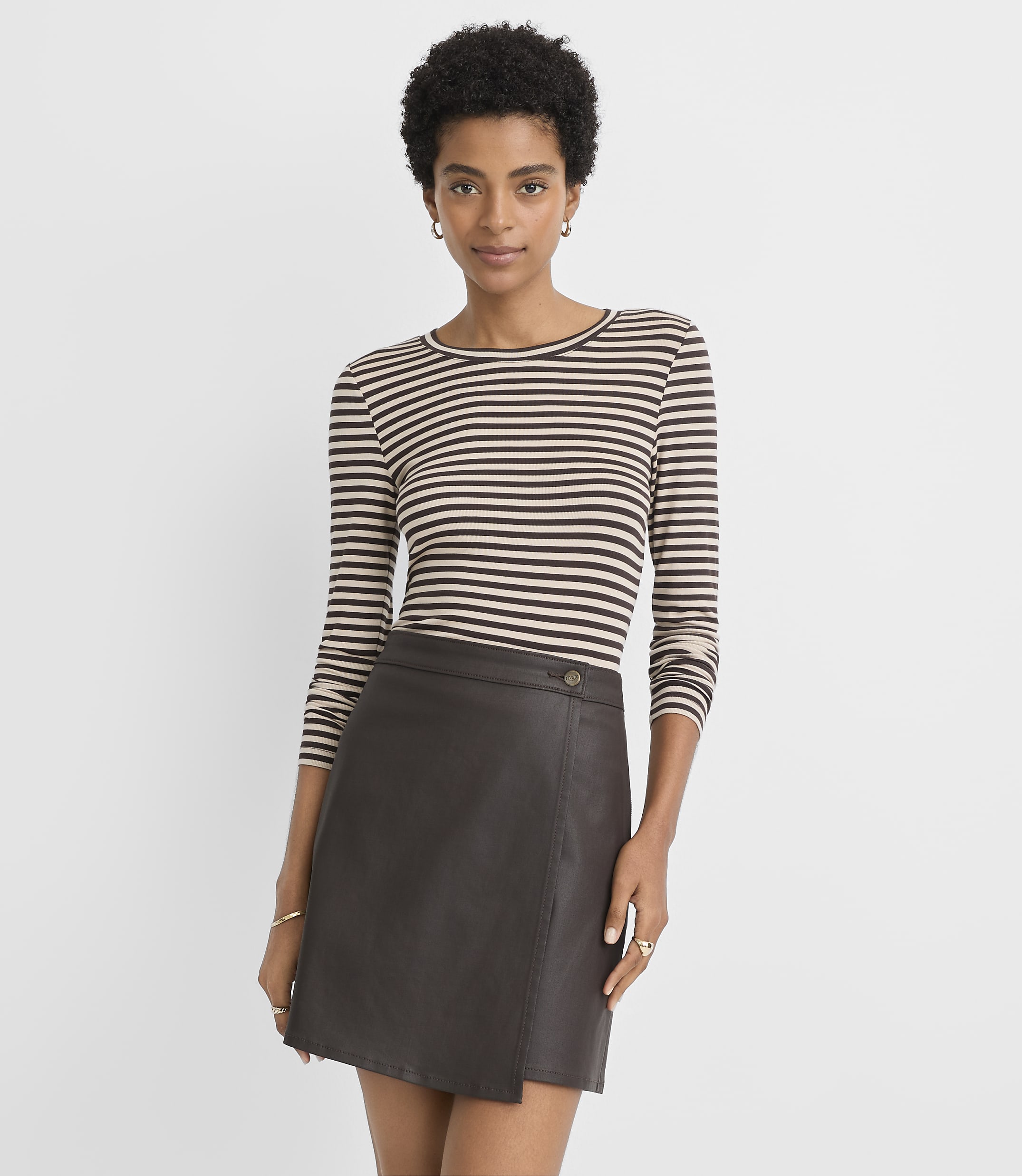 A woman in a striped shirt and a brown leather skirt stands against a plain, light gray background.