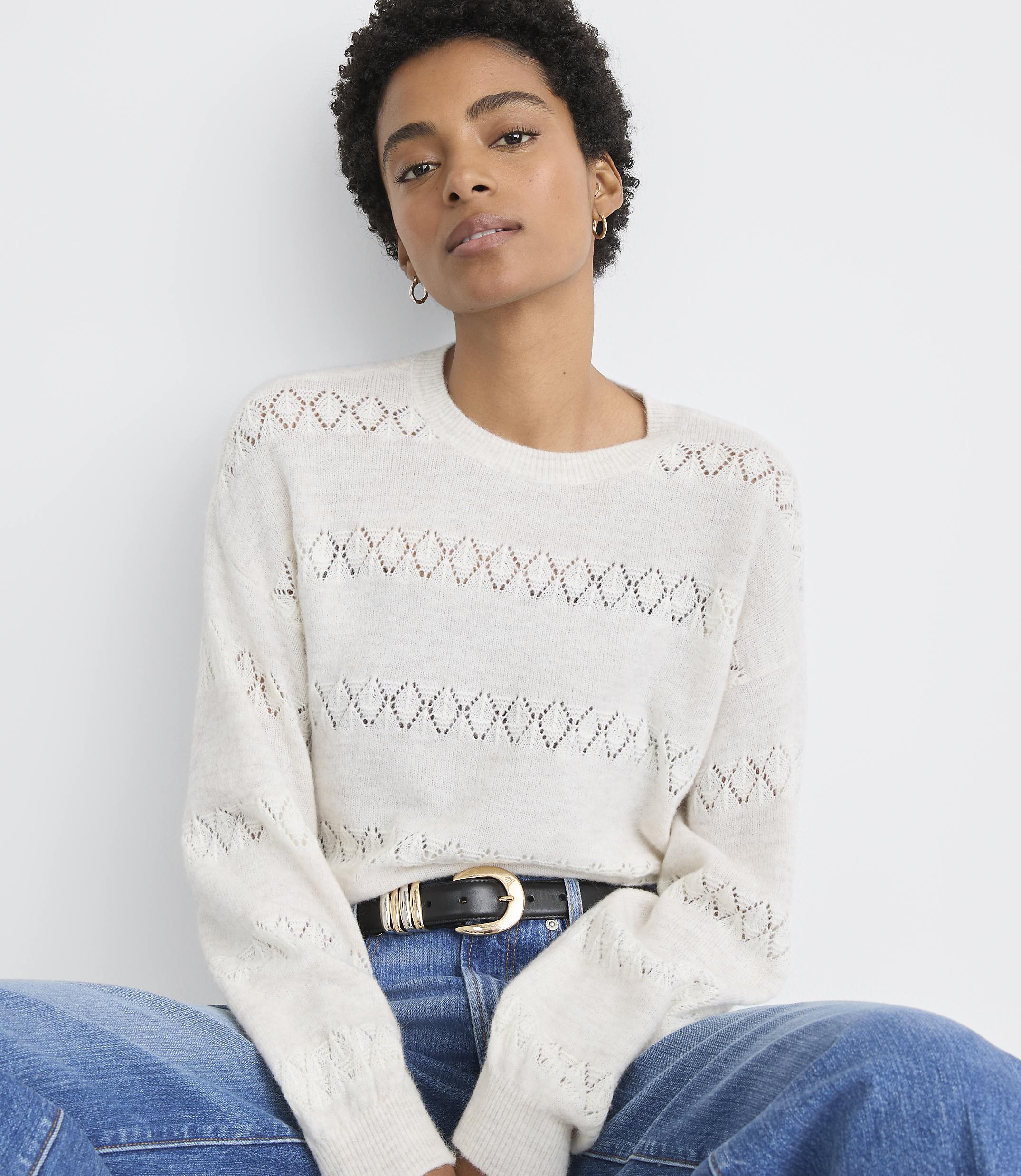 A woman in a cream-colored, open-knit sweater and blue jeans sits against a plain white background.
