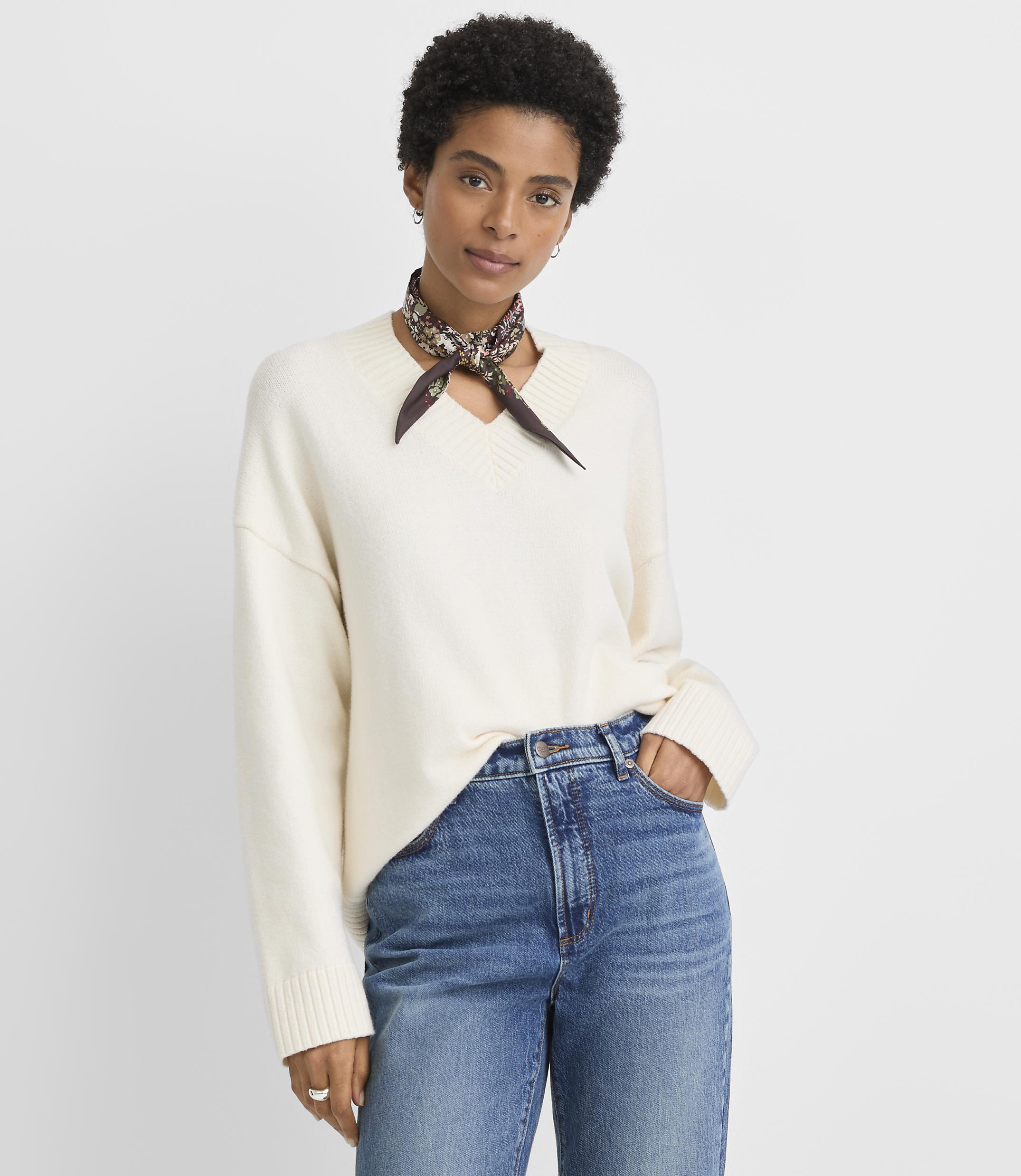 A woman in a cream v-neck sweater and blue jeans stands against a plain white background.