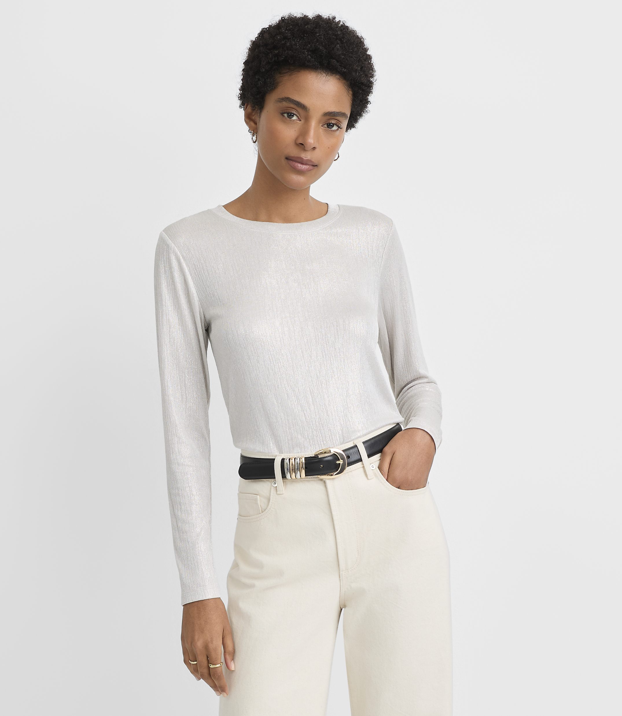 A woman in a shimmering silver long-sleeved shirt and cream pants with a black belt stands against a plain white background.