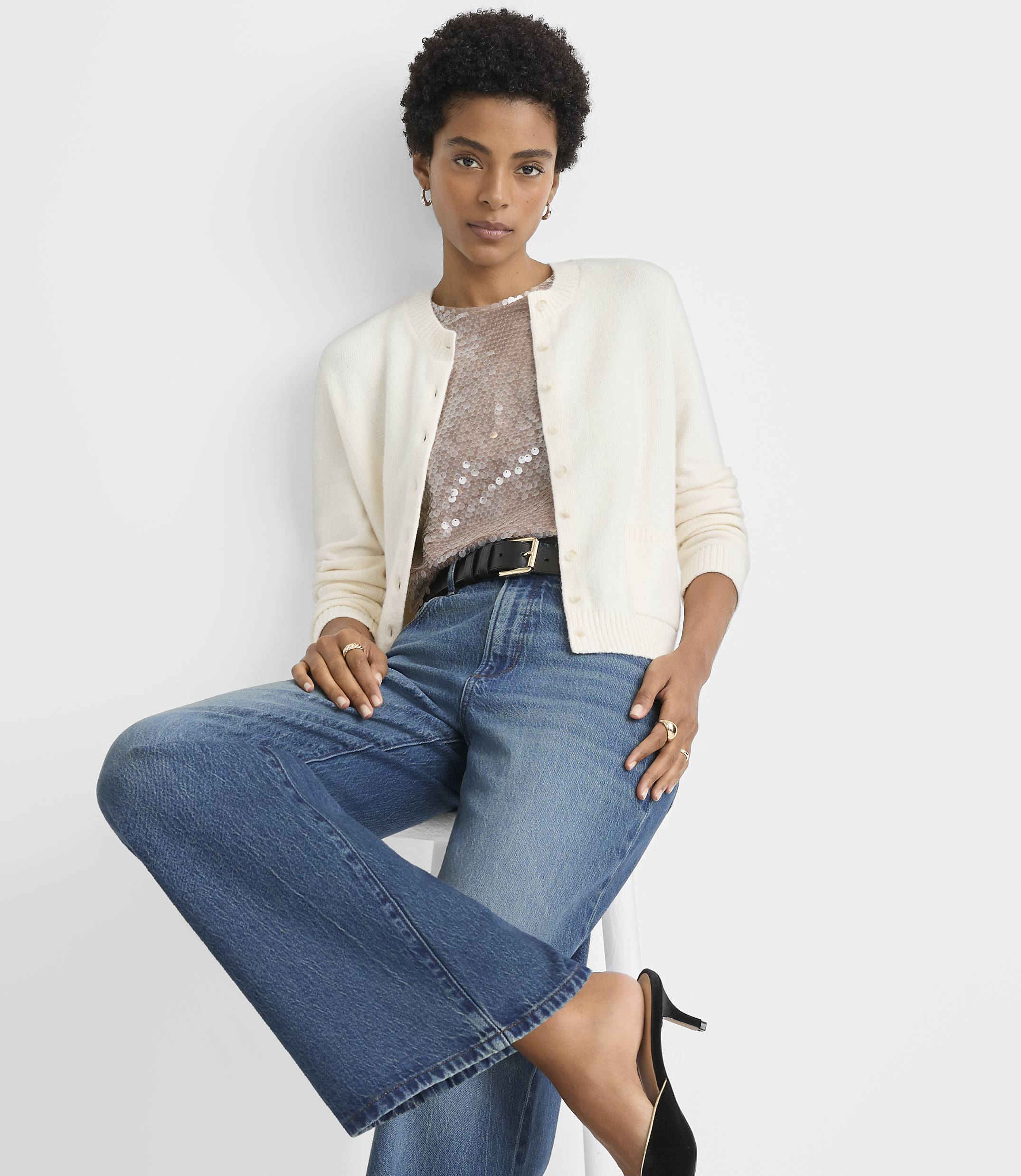 A woman in a cream cardigan, sequined top, and wide-leg jeans poses against a plain white background.