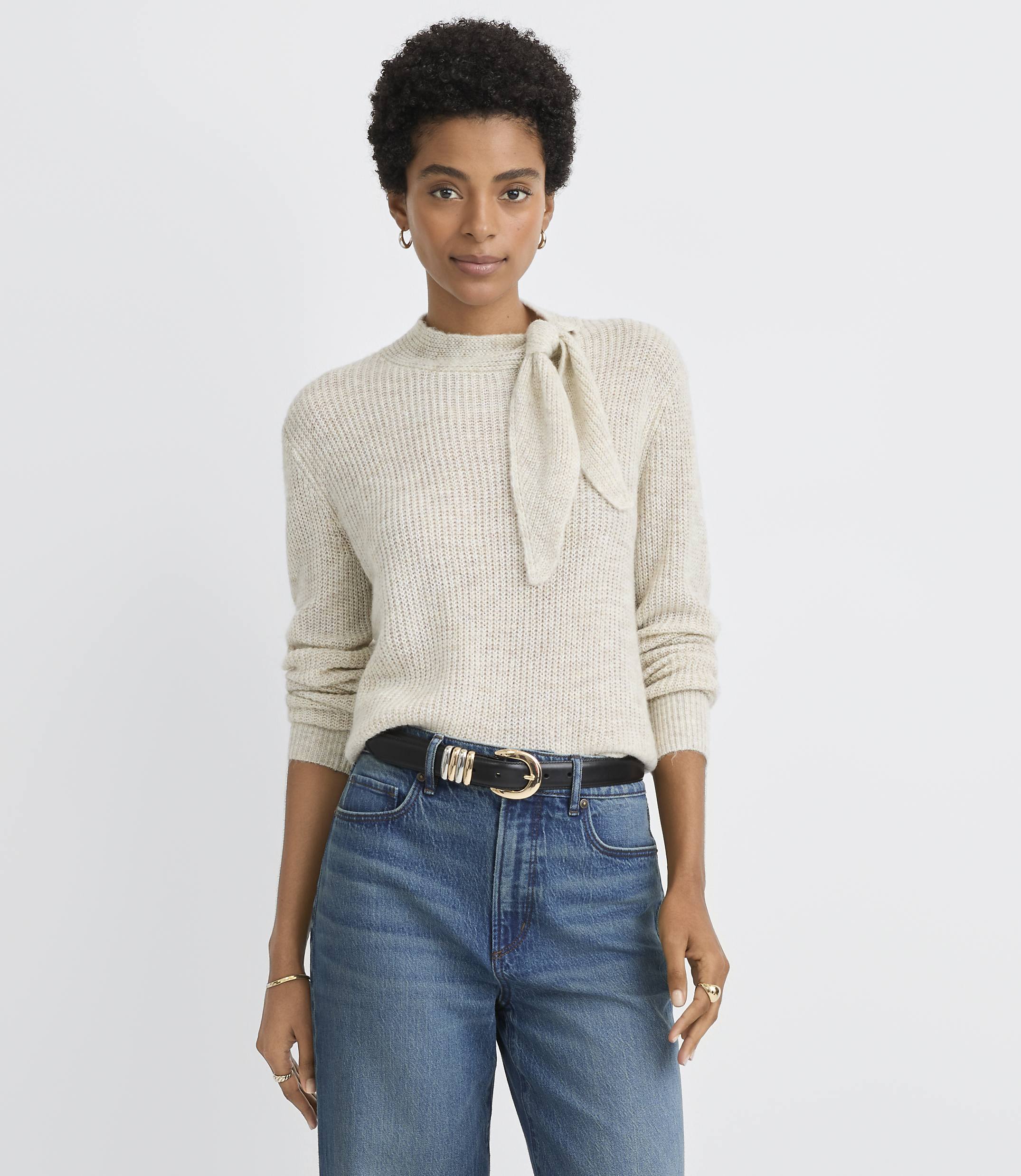 A woman in a cream-colored sweater with a bow detail and blue jeans stands against a plain, light gray background.