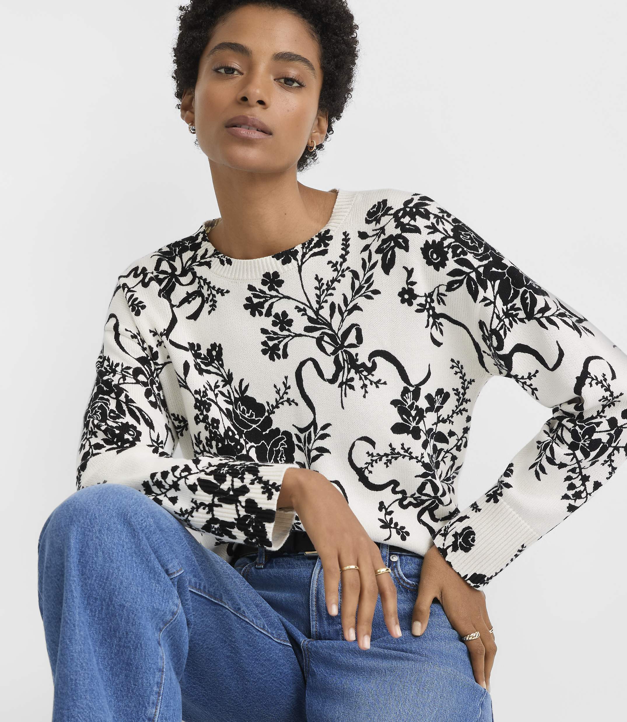 A woman in a black and white floral sweater and blue jeans is shown against a plain white background.
