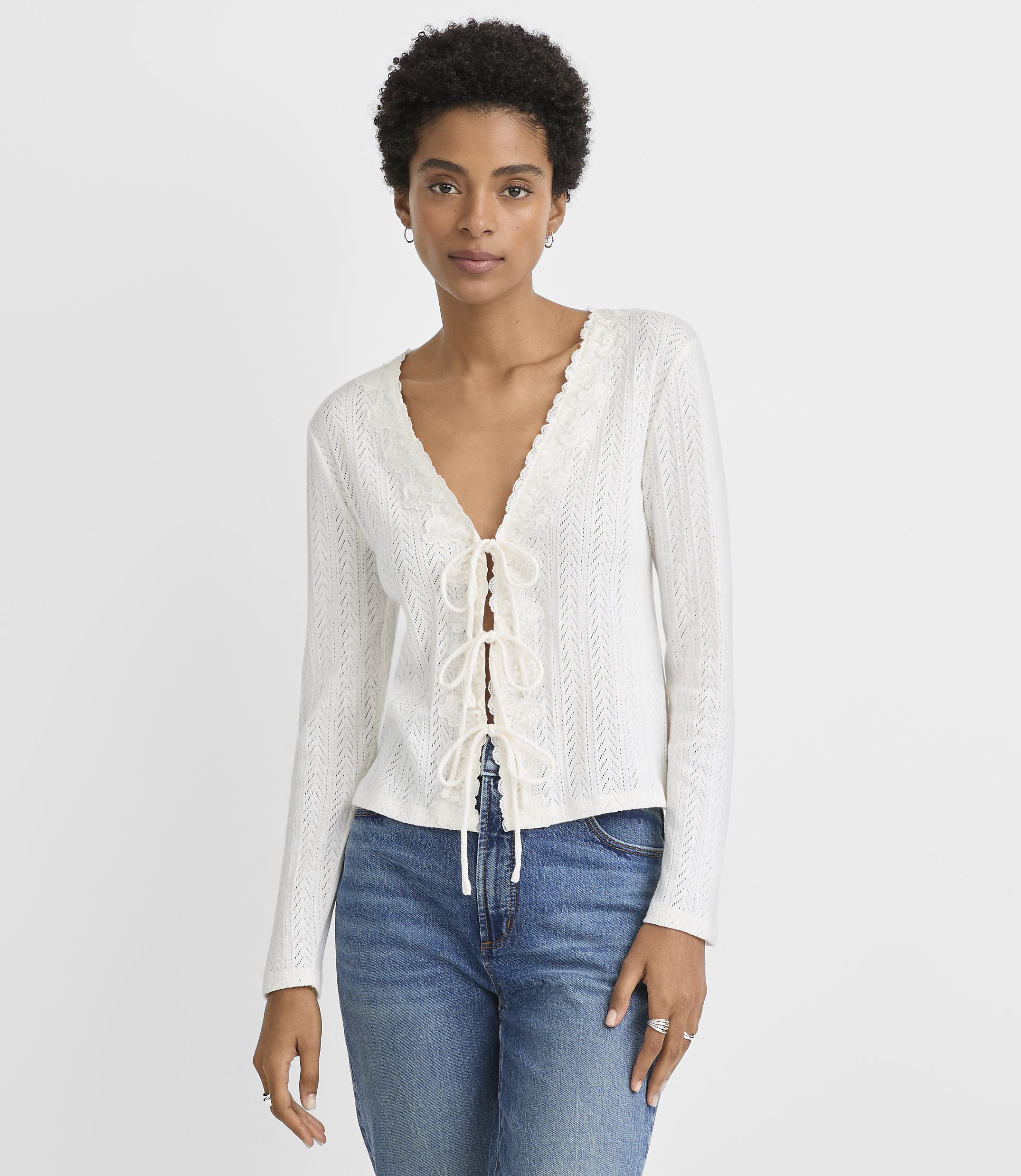A woman in a white lace-up top and blue jeans stands against a plain white background.