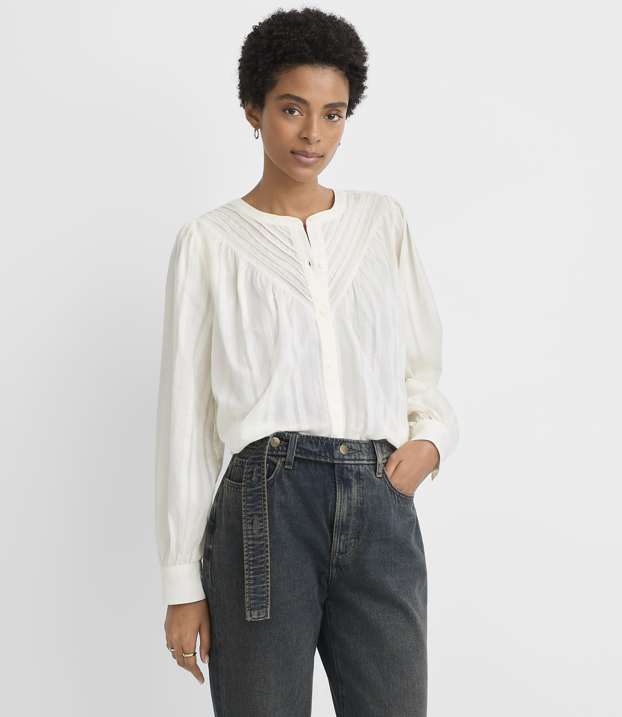 A woman in a white blouse and dark jeans stands against a plain white background.
