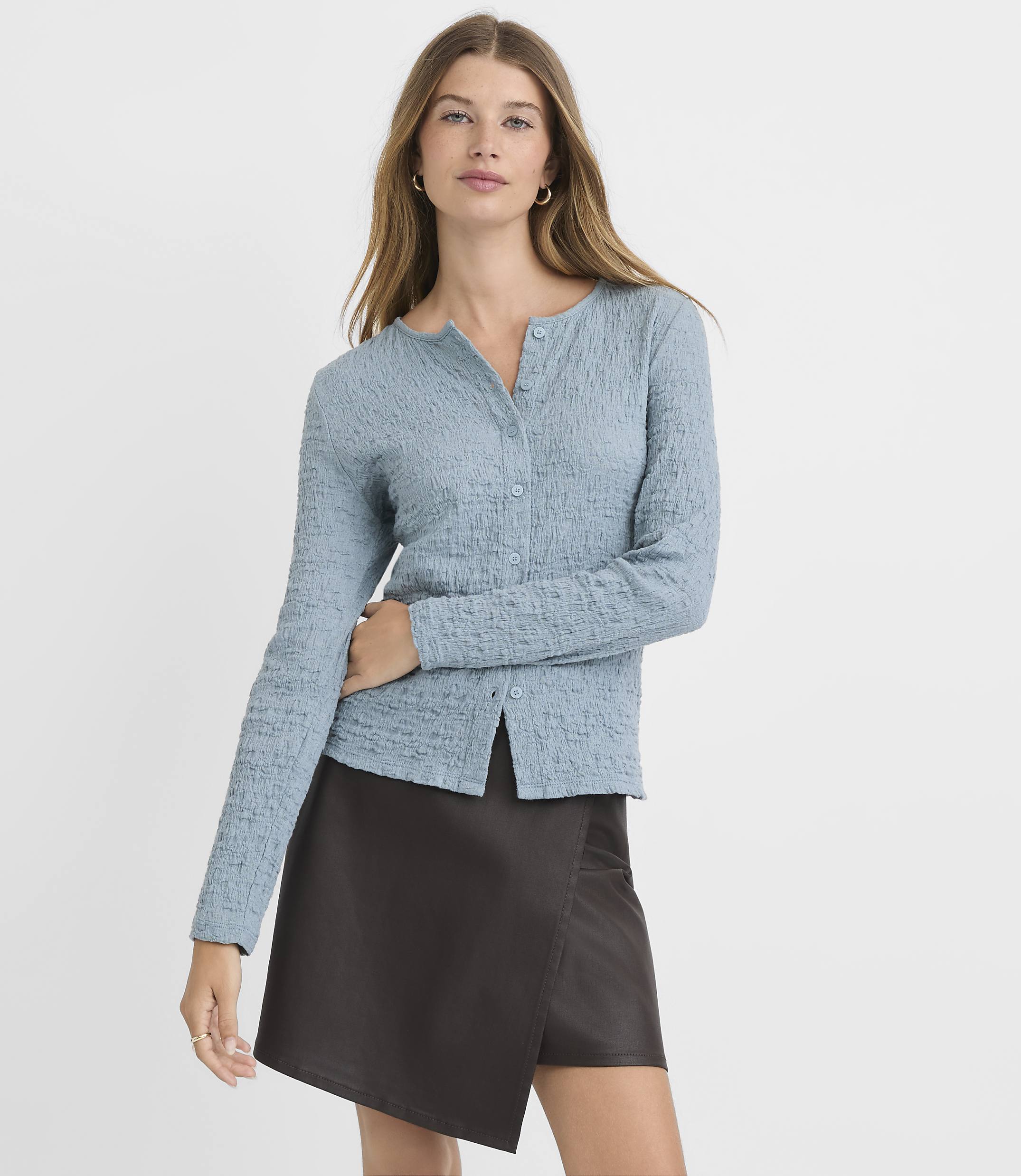 A woman in a textured blue cardigan and a dark brown skirt stands against a plain, light gray background.