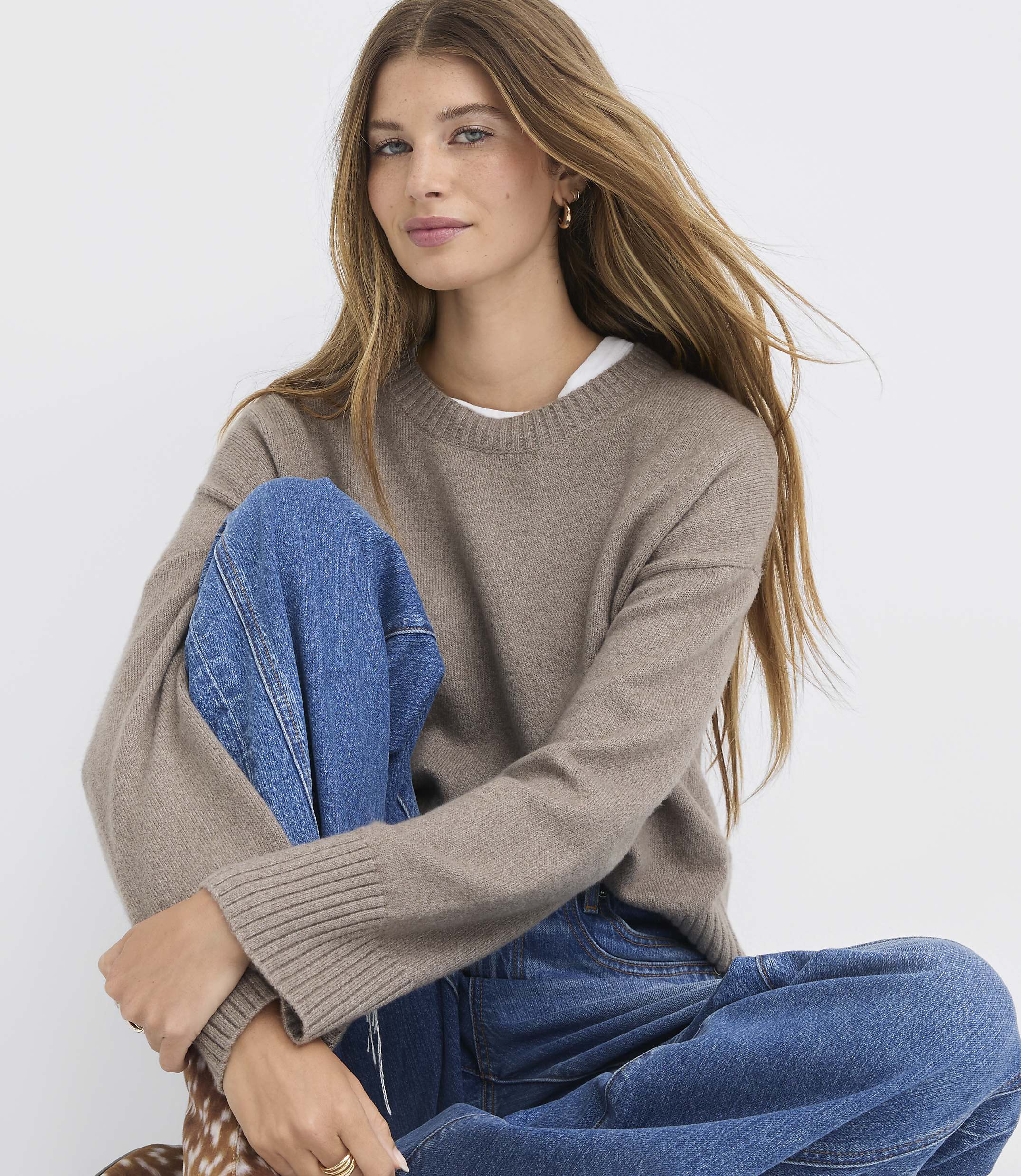 A young woman with long, blonde hair, wearing a brown sweater and blue jeans, sits against a plain white background.