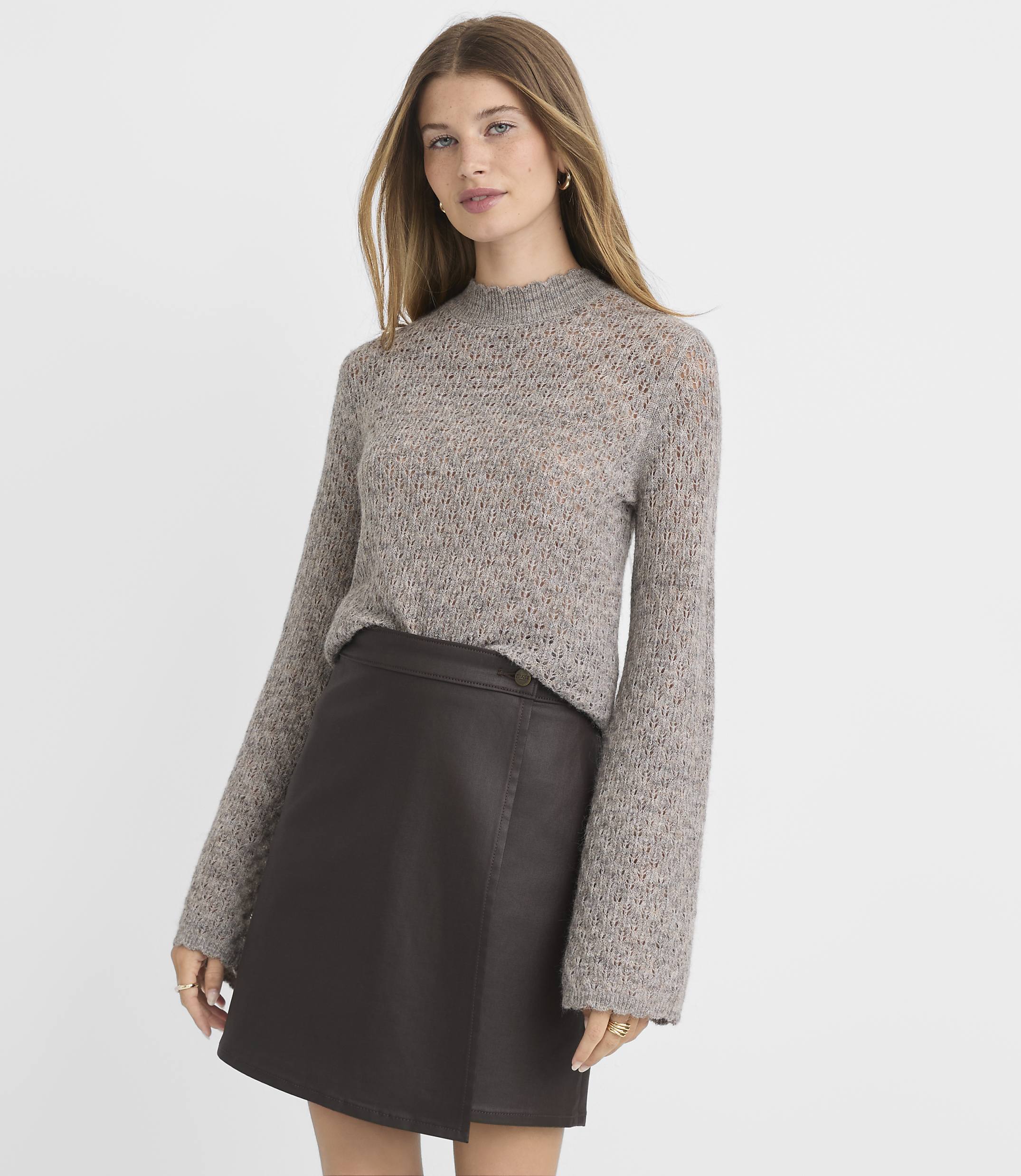 A woman in a grey knit sweater and a dark brown faux leather skirt stands against a plain white background.