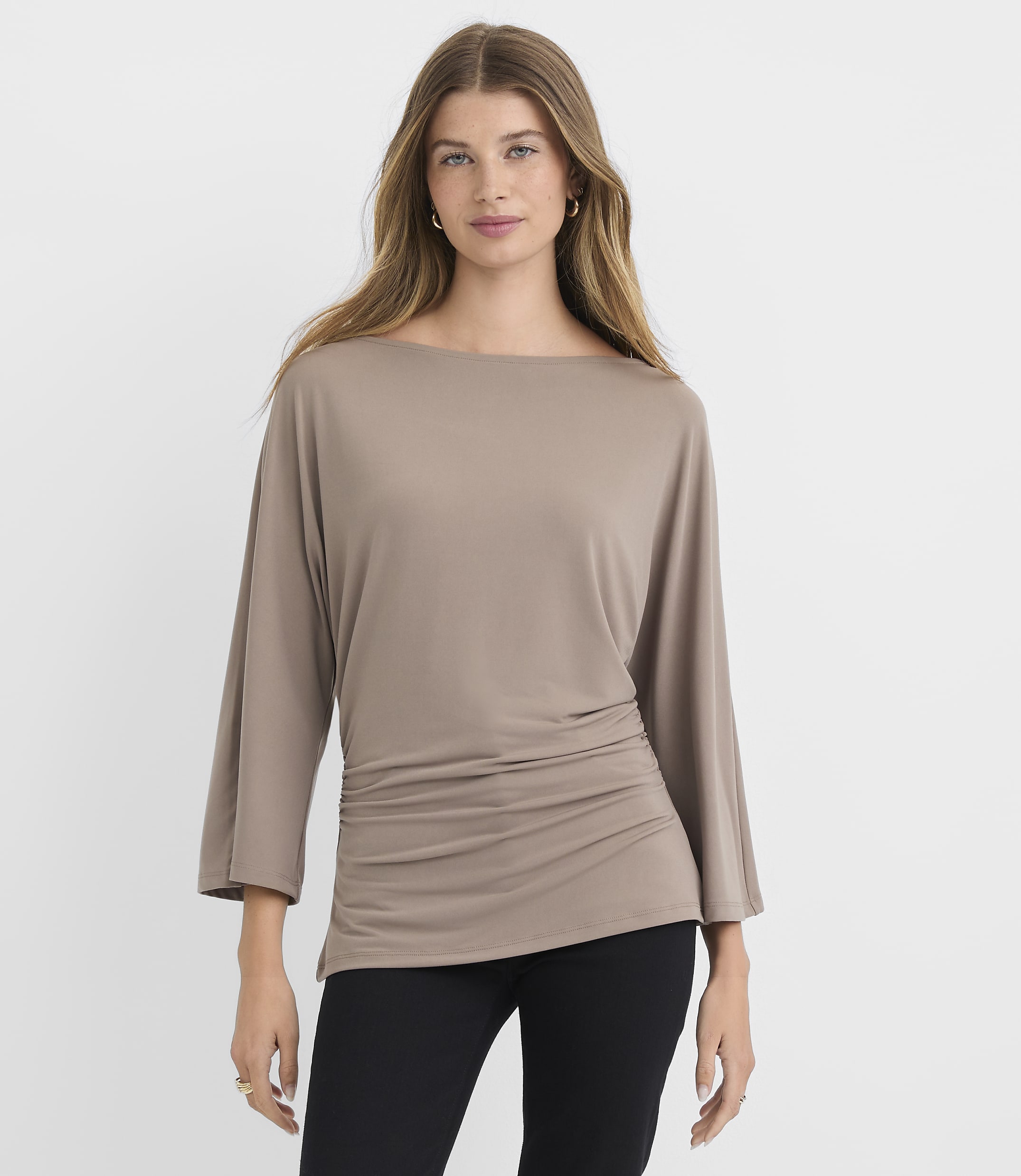 A woman in a taupe dolman-sleeve top and black pants stands against a plain white background.