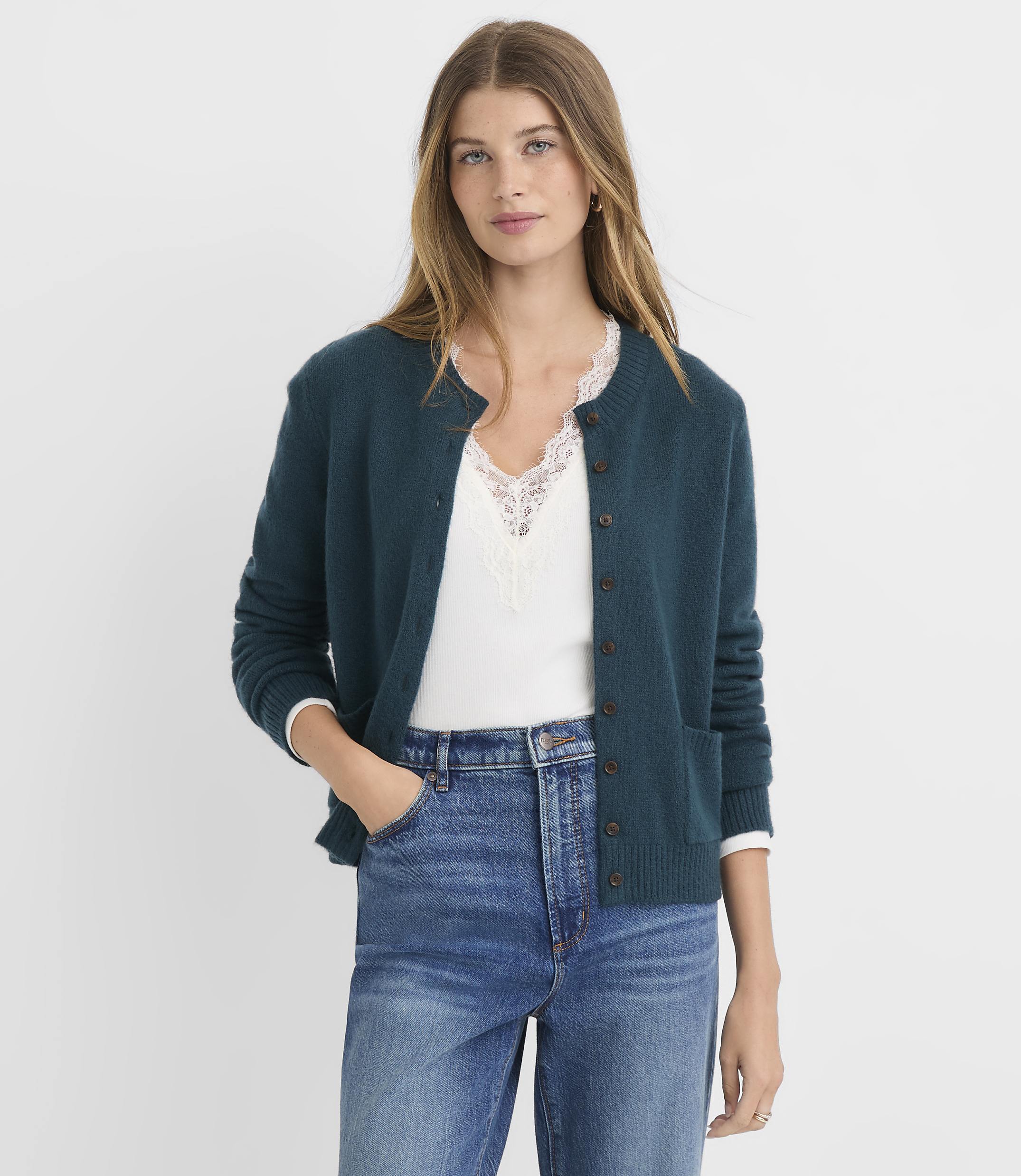 A woman in a teal cardigan and blue jeans stands against a plain white background.