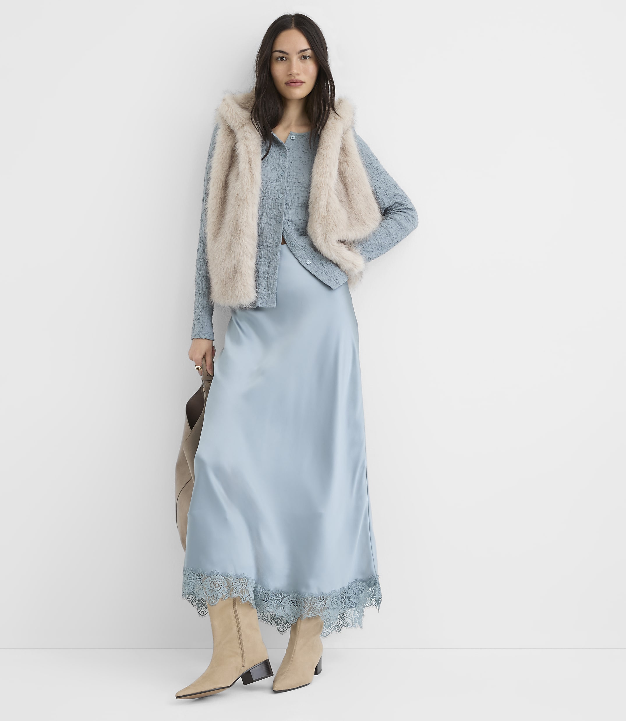 A woman in a light blue satin slip dress and a fuzzy vest stands against a plain white background.