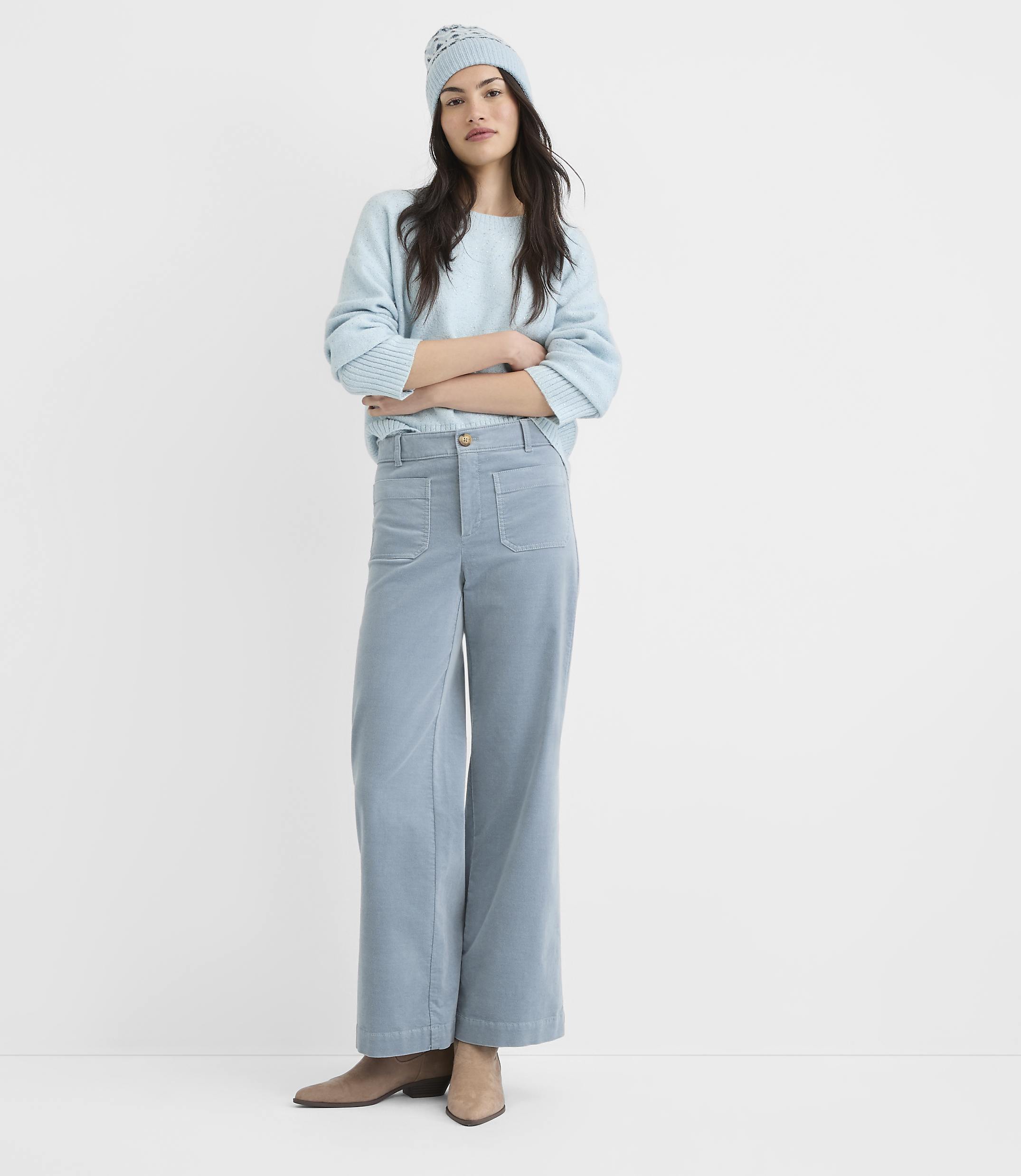 A woman in a light blue beanie, sweatshirt, and corduroy pants stands with her arms crossed against a plain white background.