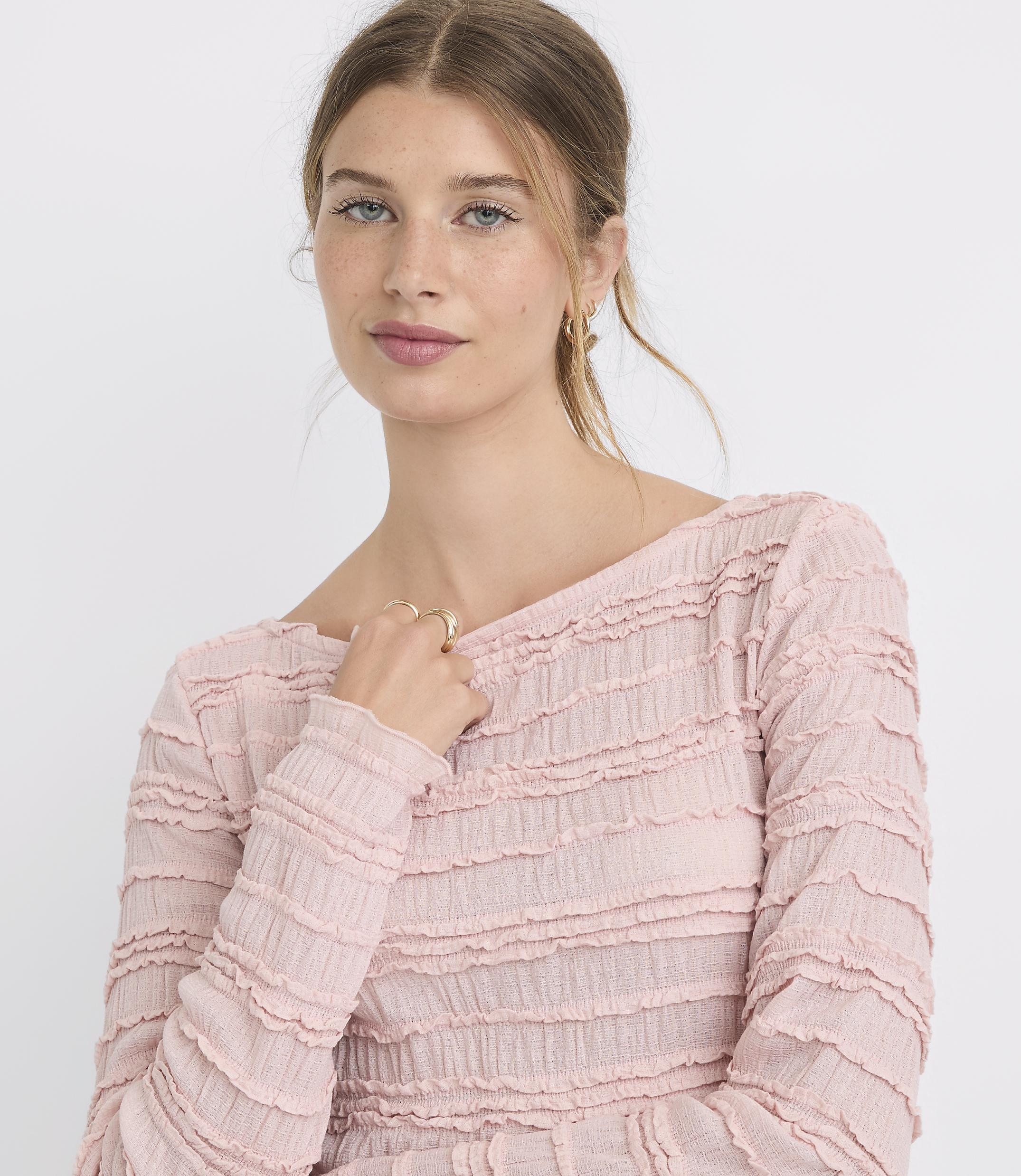 A young woman with blonde hair pulled back wears a textured pink top, with a plain white background.
