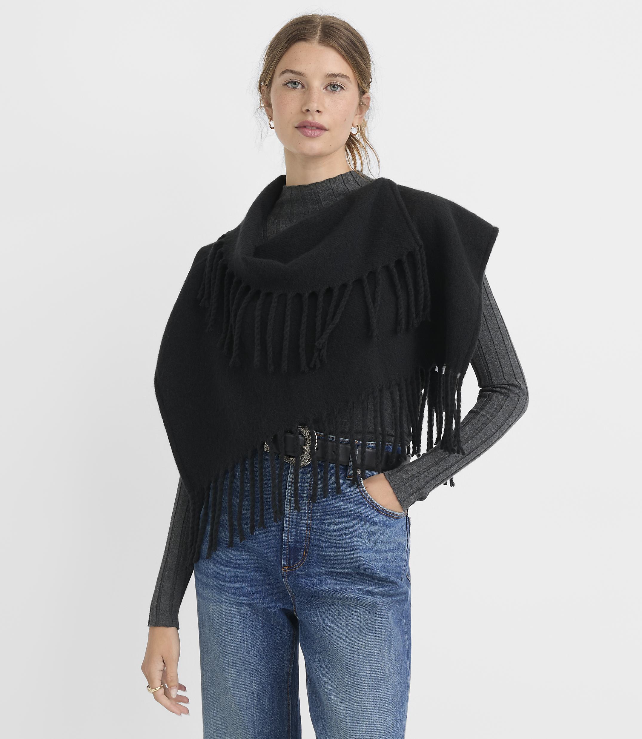 A woman in a black fringed shawl and jeans stands against a plain gray background.