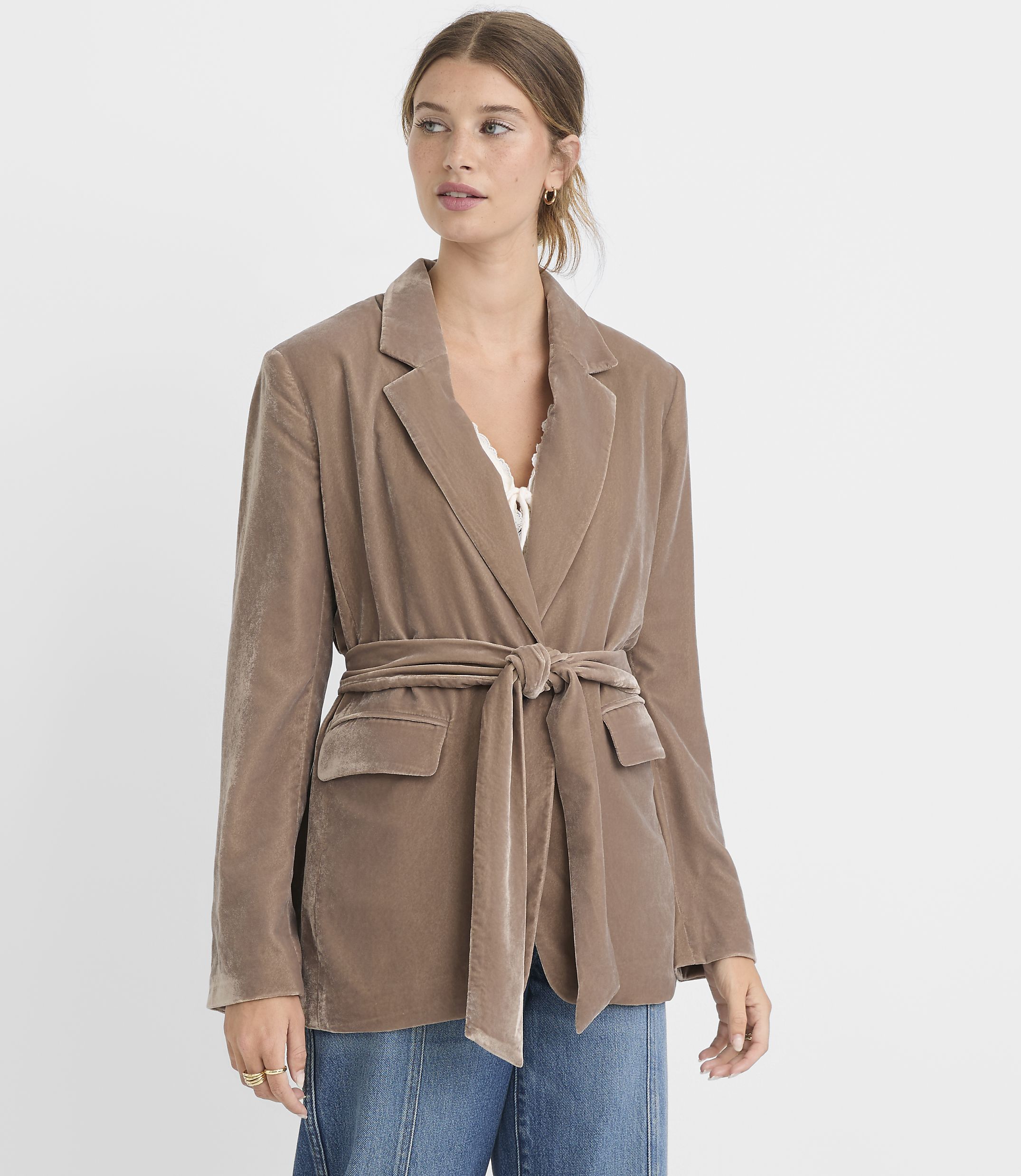 A woman in a taupe velvet blazer cinched with a matching belt stands against a plain white background.