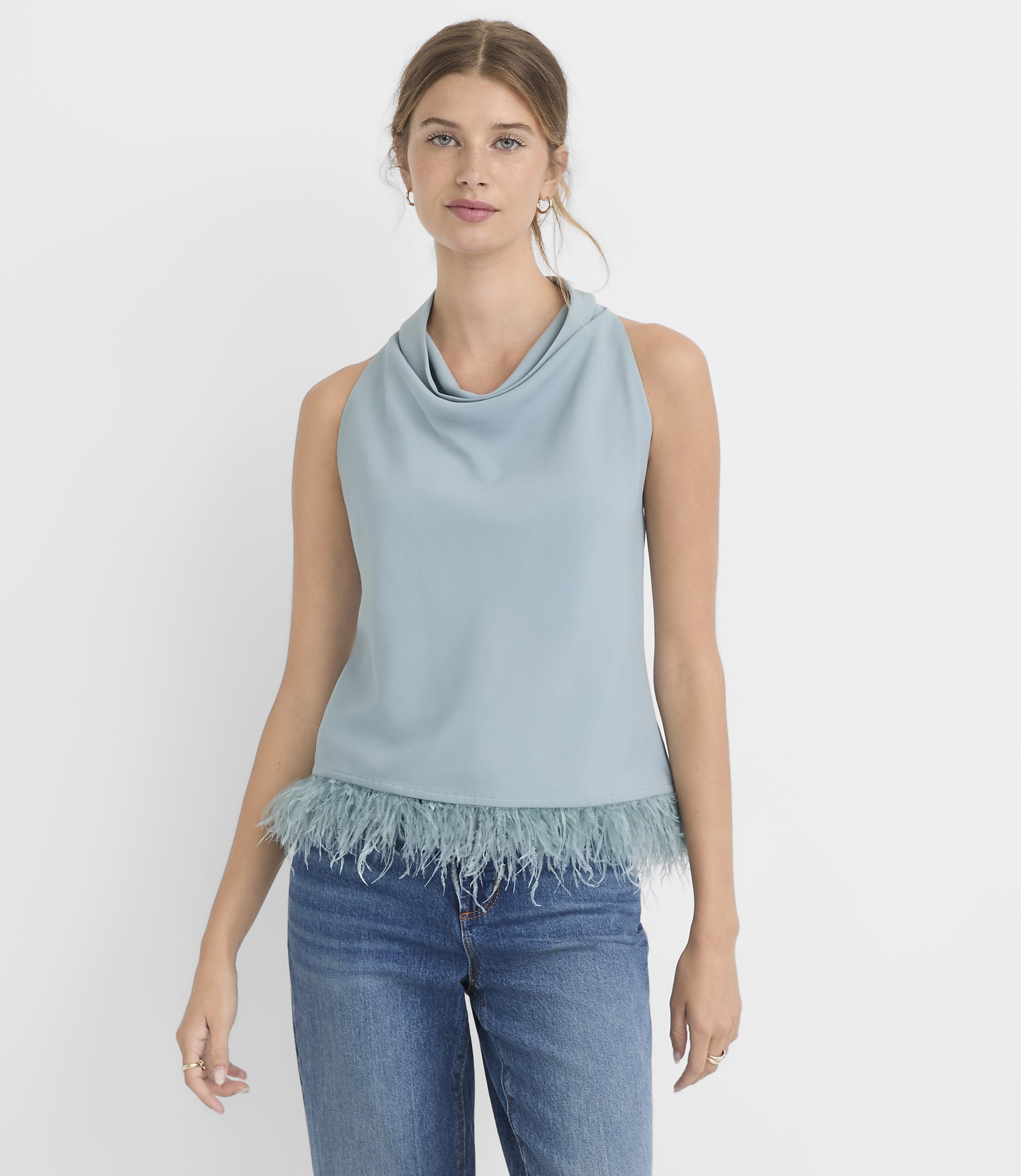 A woman in a blue cowl-neck sleeveless top with a feathered hem and blue jeans stands against a plain white background.