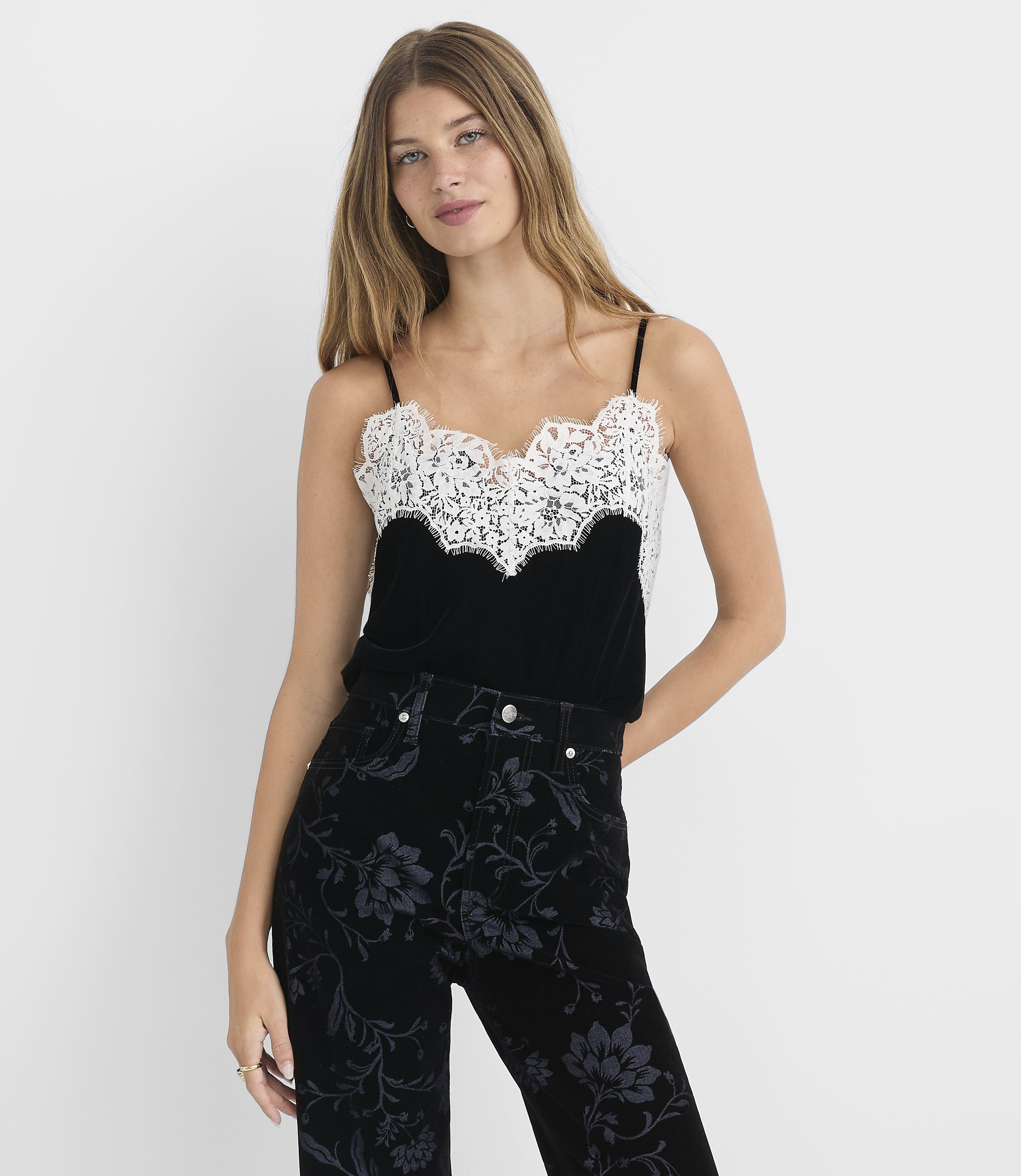 A woman in a black velvet camisole with white lace trim and patterned black pants stands against a plain white background.