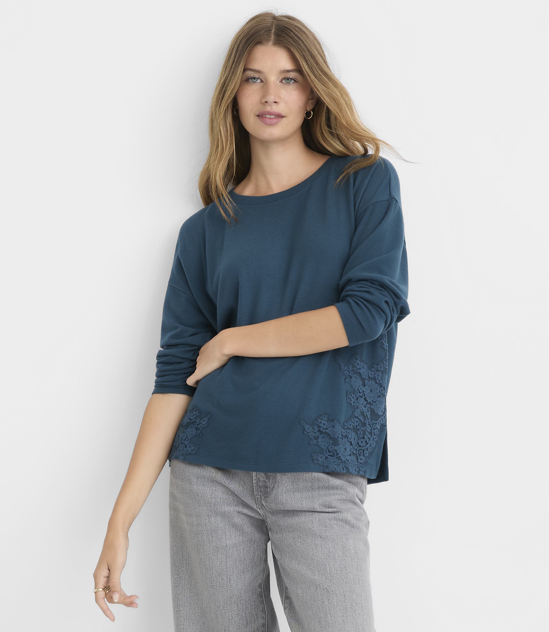 A woman in a teal long-sleeved shirt with lace detailing on the sides stands against a plain gray background.
