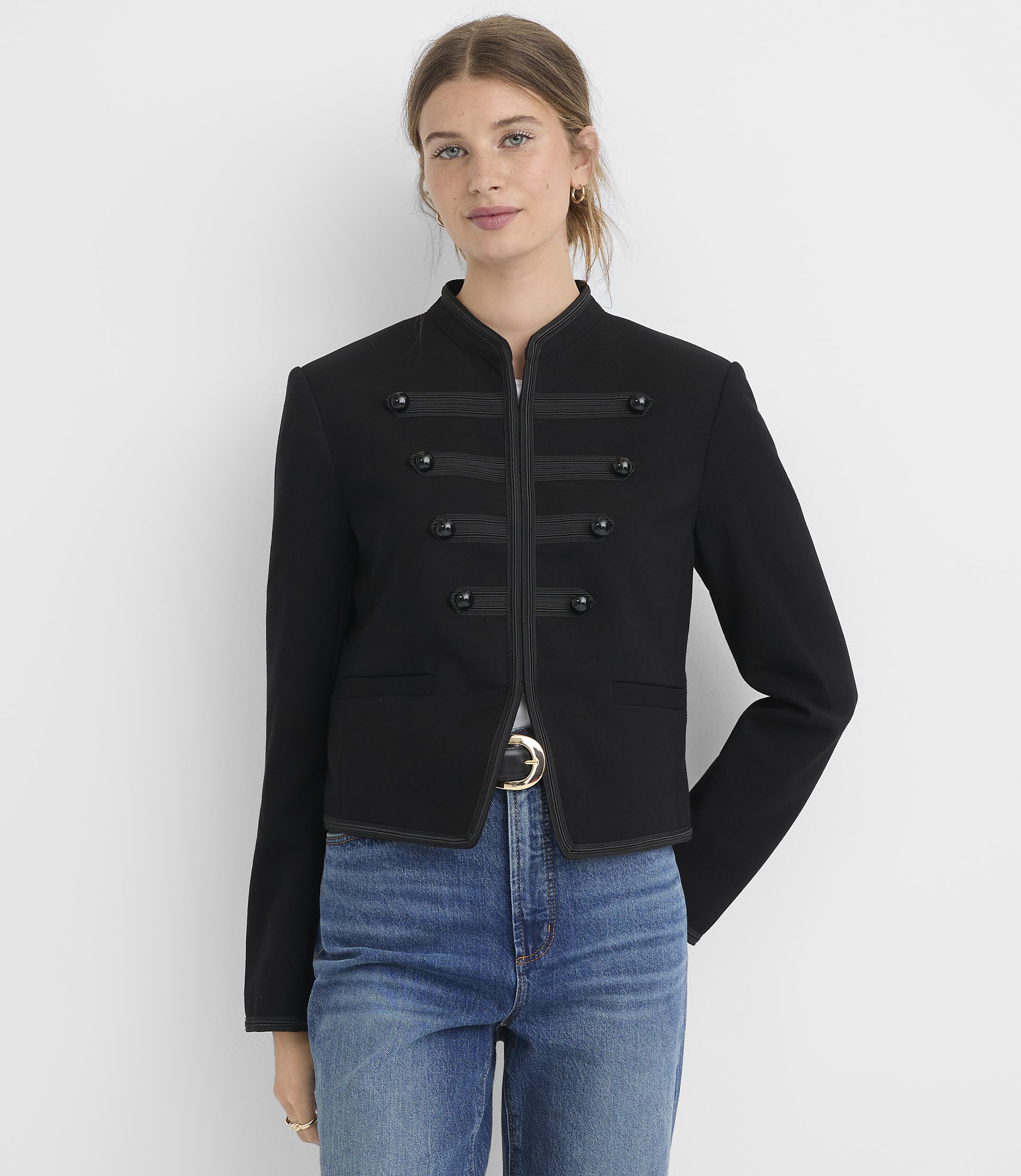 A woman in a black military-style jacket and blue jeans stands against a plain white background.