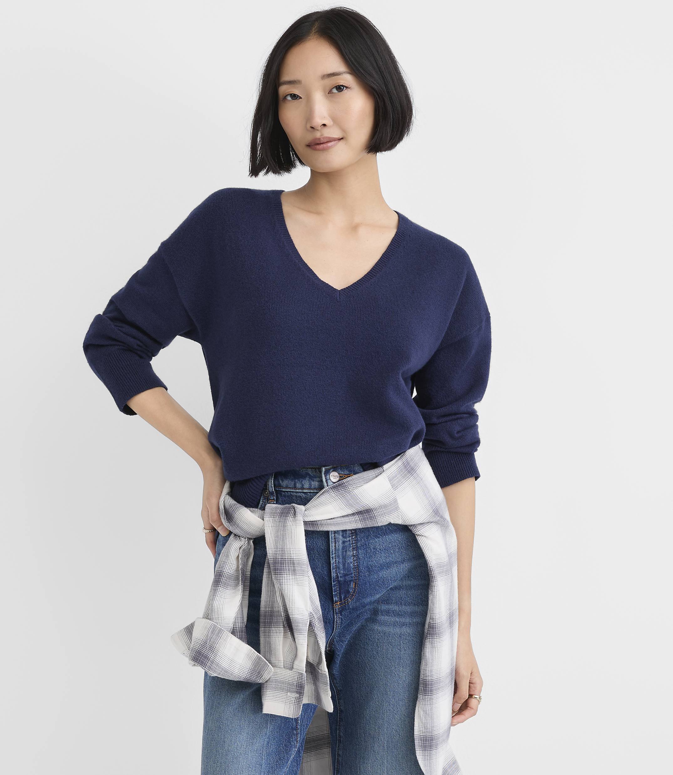 A woman wearing a navy blue V-neck sweater and blue jeans with a plaid shirt tied around her waist stands against a plain white background.