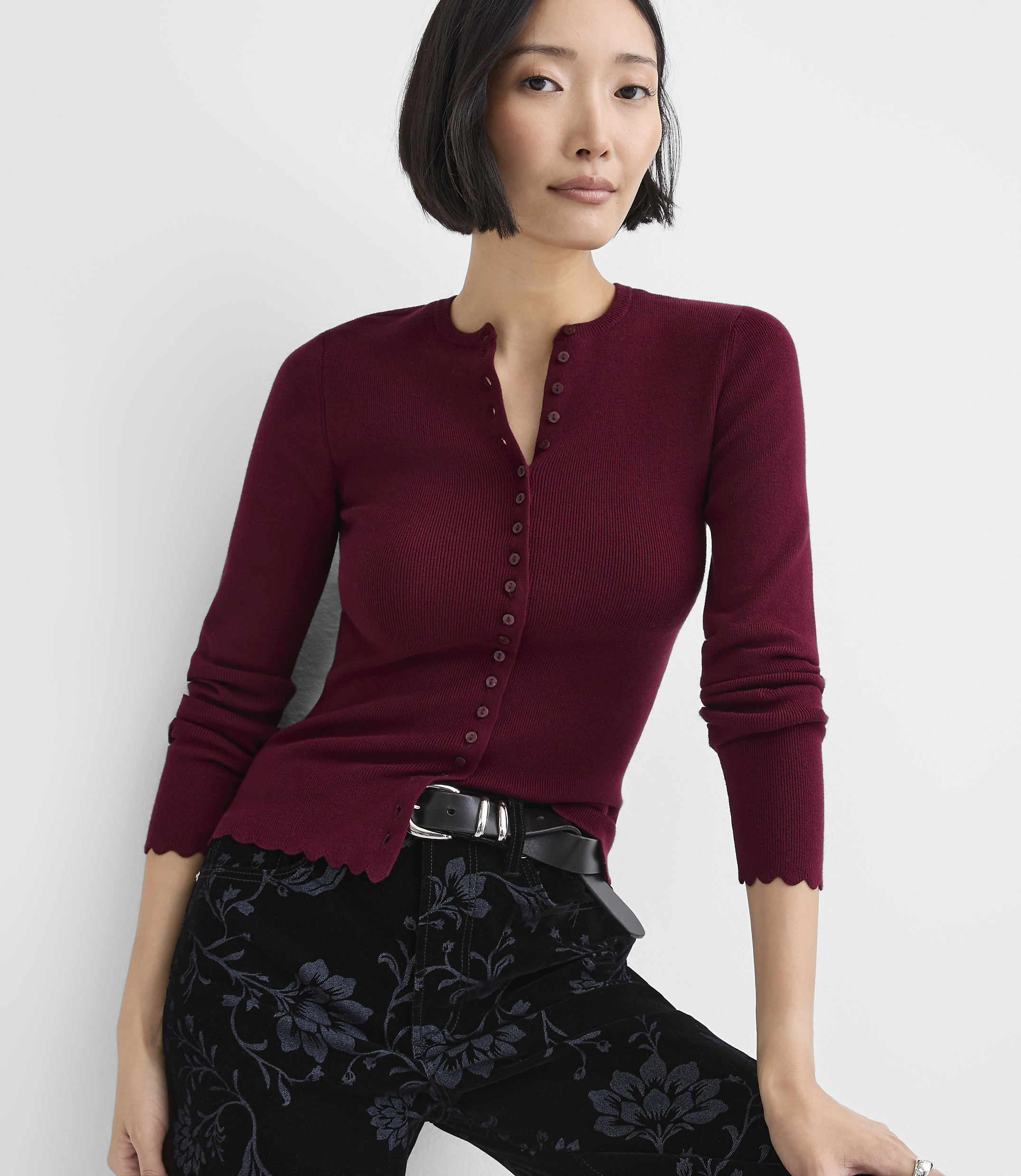 A woman wearing a burgundy button-down cardigan and floral black trousers poses against a plain white background.