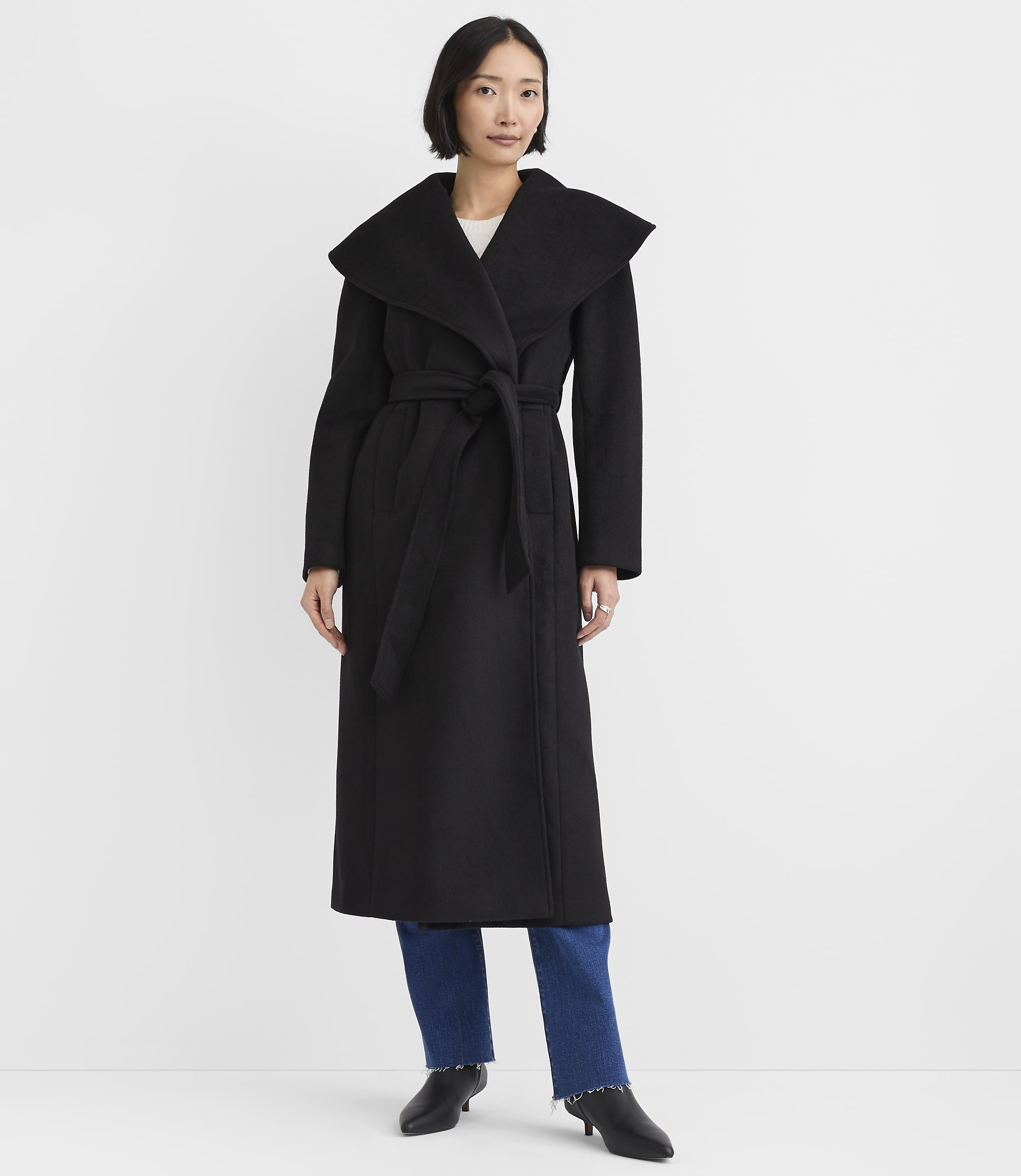 A woman wearing a long black belted coat and blue jeans stands against a plain white background.