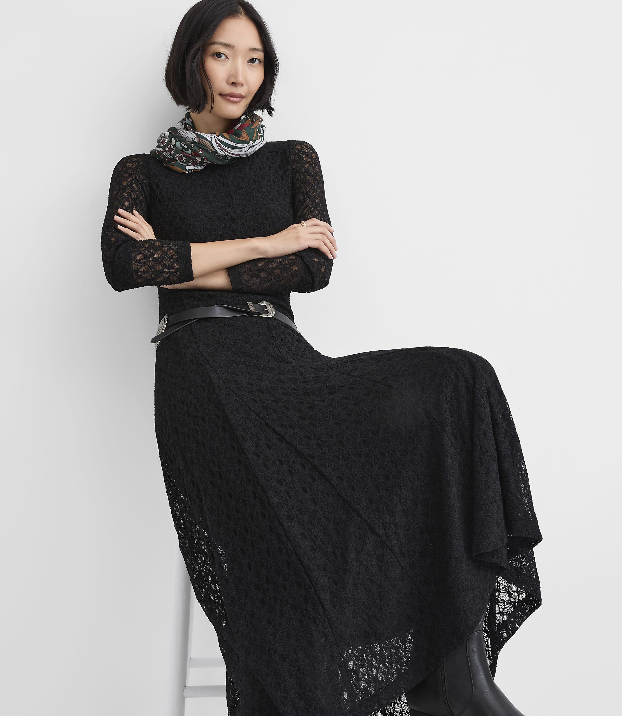 A woman wearing a black lace dress and a patterned scarf poses against a plain white background.