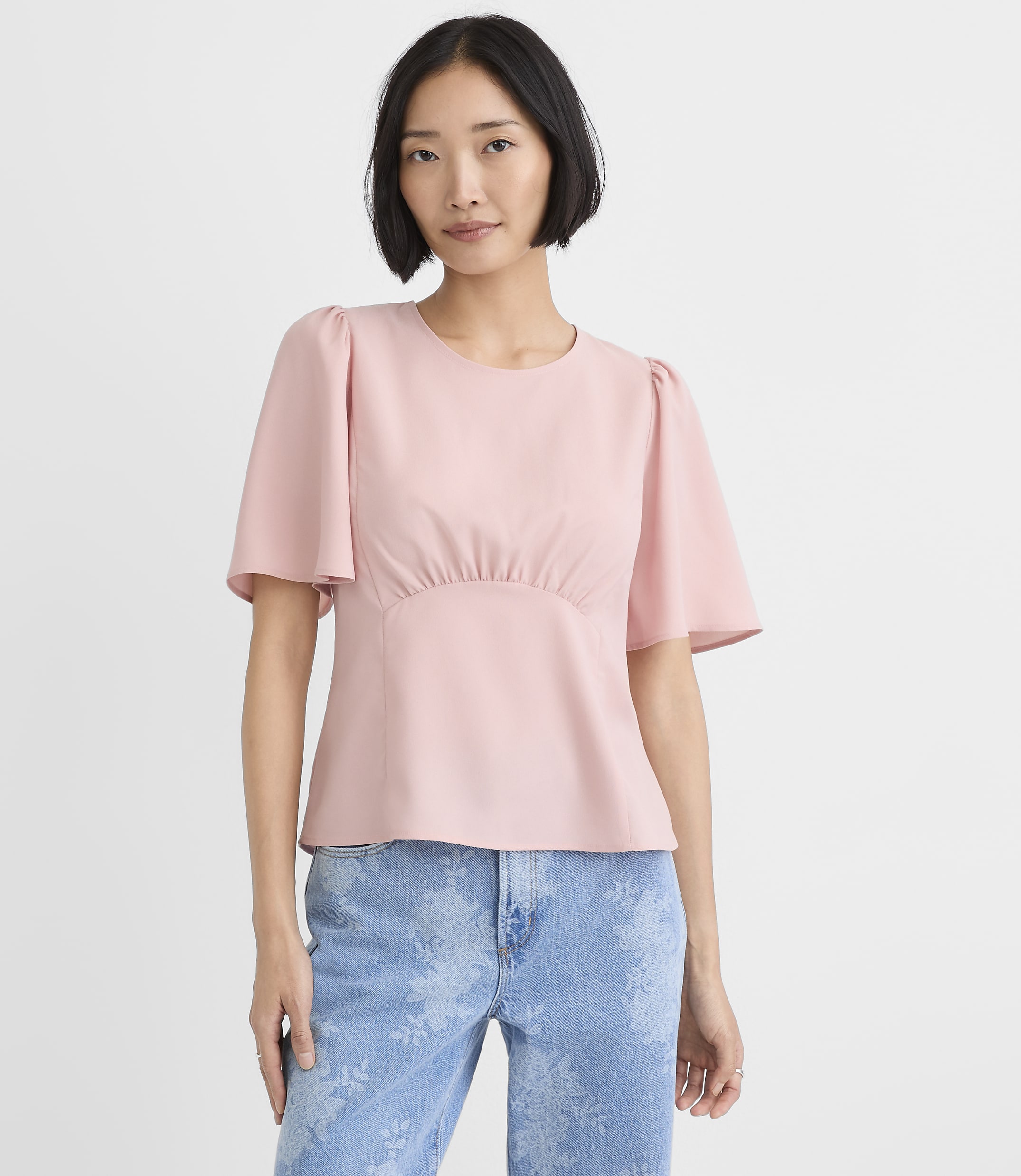 A woman wearing a light pink short-sleeved top and blue patterned jeans stands against a plain white background.