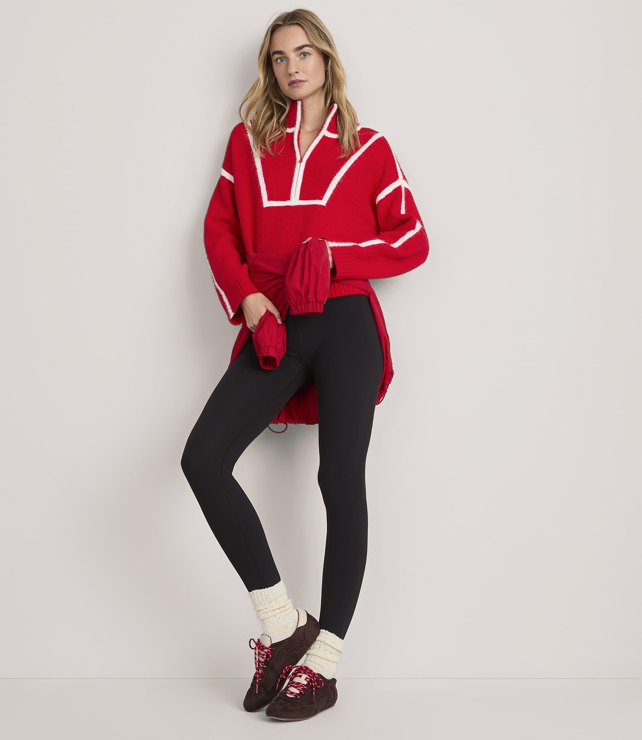 A woman wearing a red oversized sweater, black leggings, and brown boots stands against a plain white background.