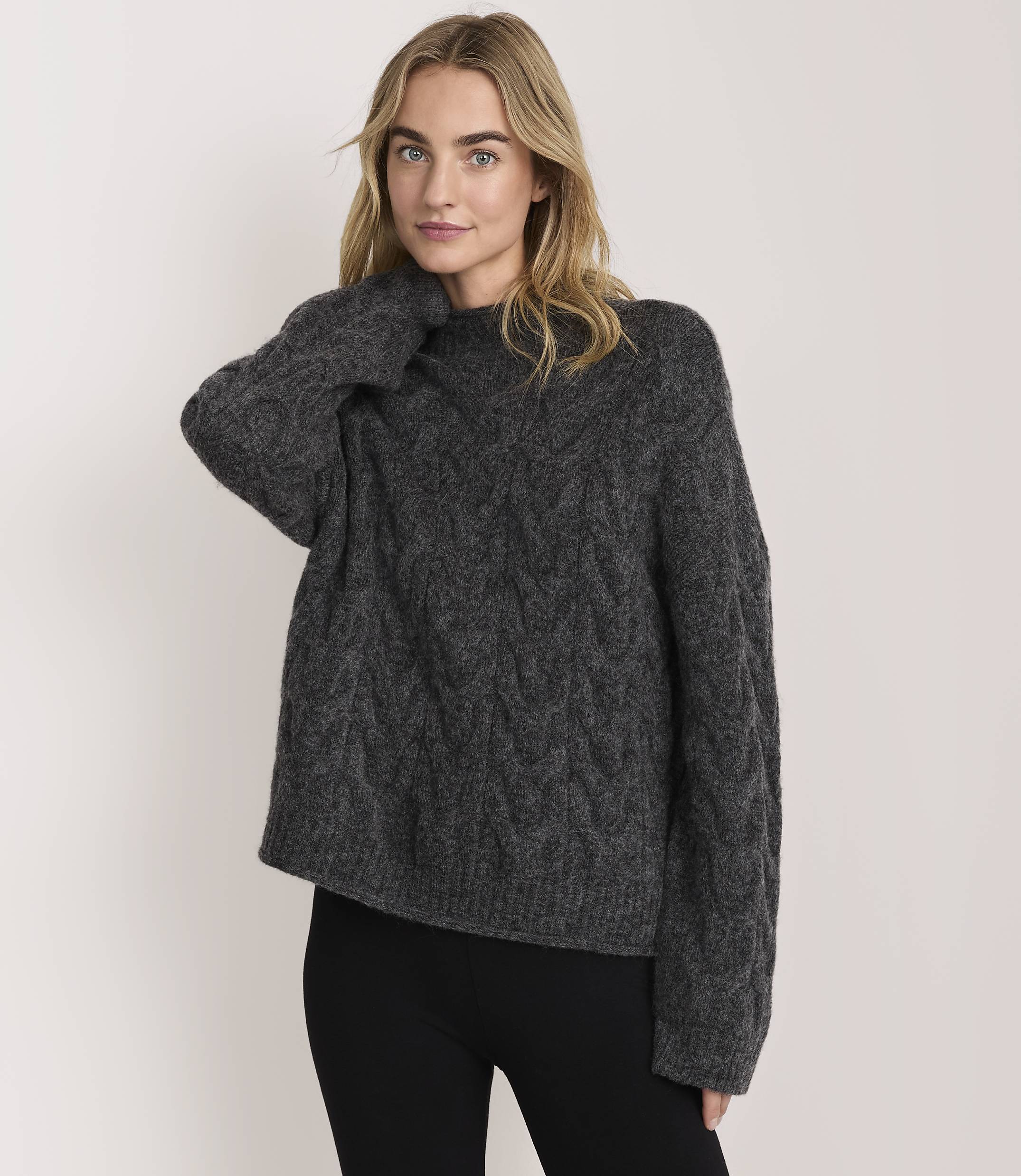 A blonde woman wearing a charcoal grey cable-knit sweater and black leggings stands against a plain white background.