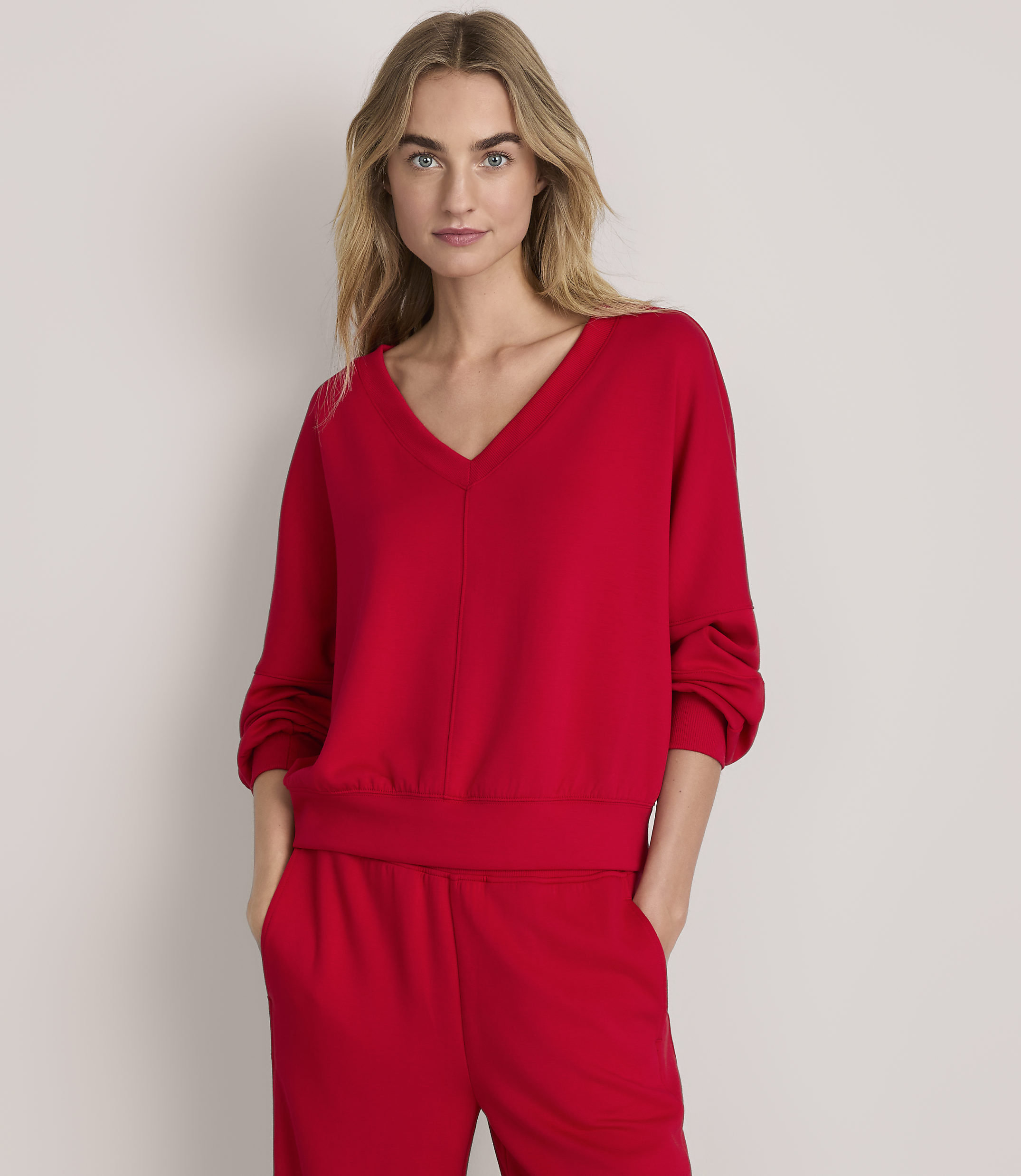 A woman wearing a red v-neck sweatshirt and matching red pants stands against a plain, light-colored background.
