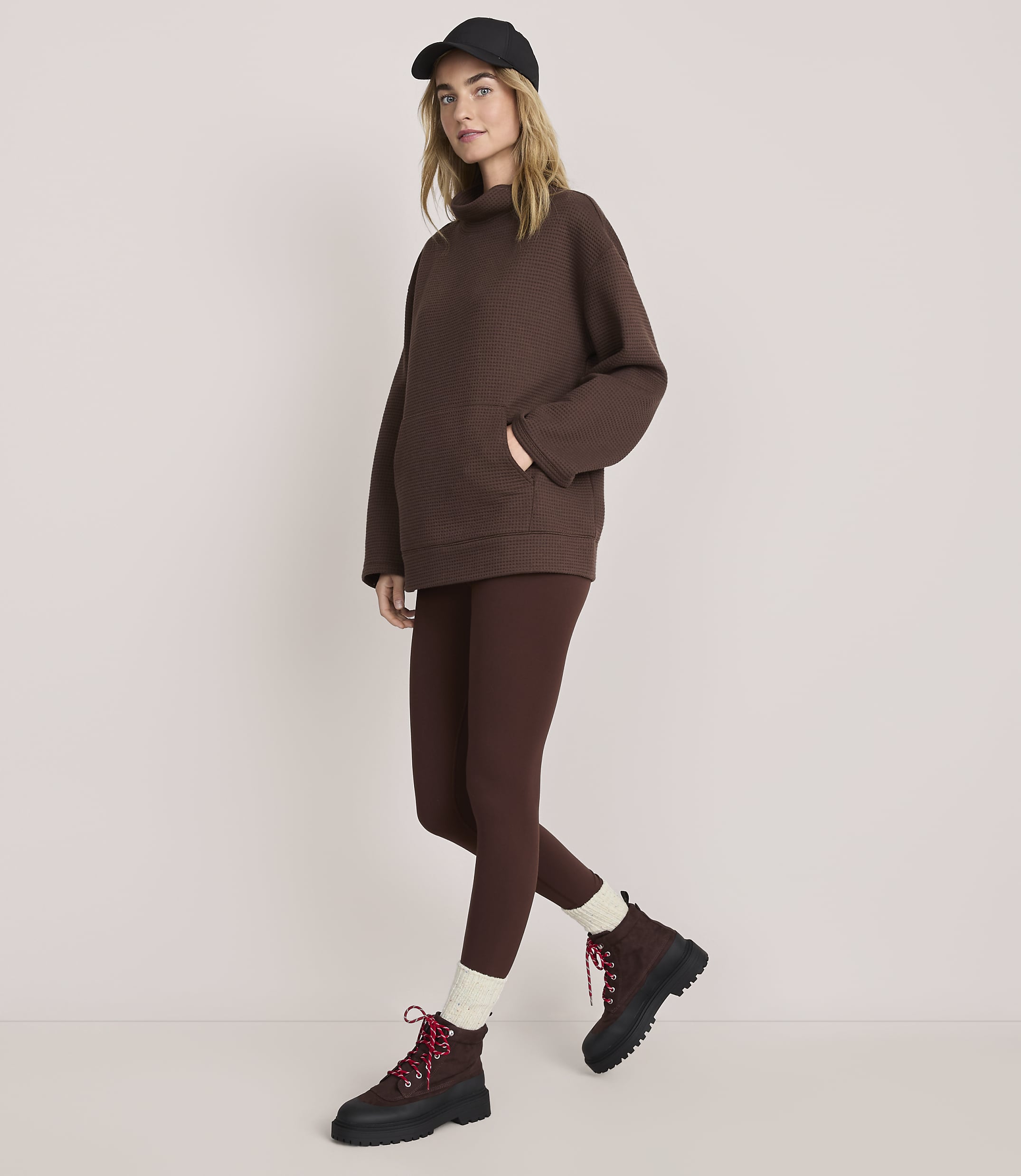 A woman wearing a brown oversized sweater, matching leggings, a black cap, and brown boots stands against a plain, light-colored background.