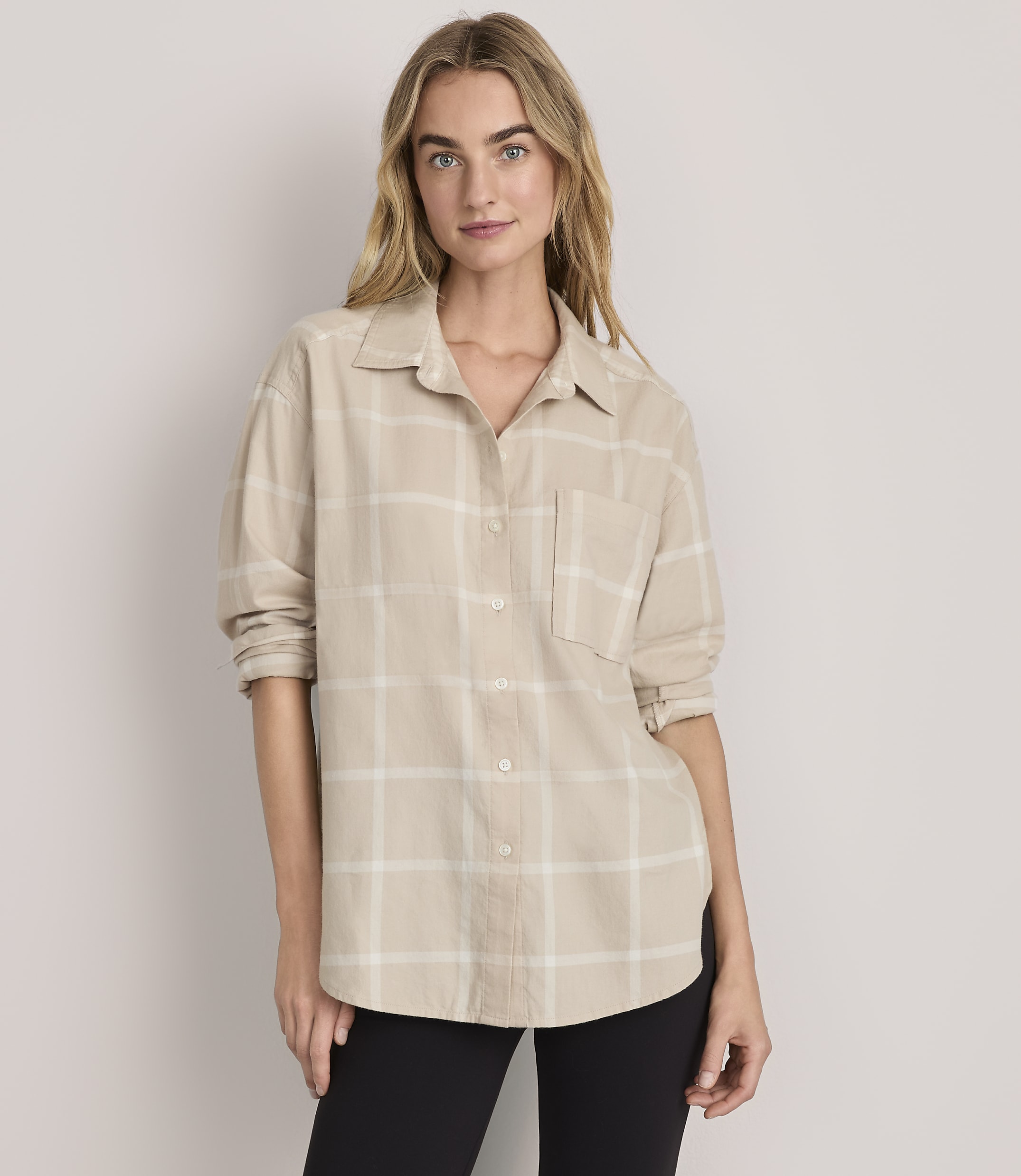 A woman wearing a beige plaid button-down shirt and black pants stands against a plain, light-colored background.
