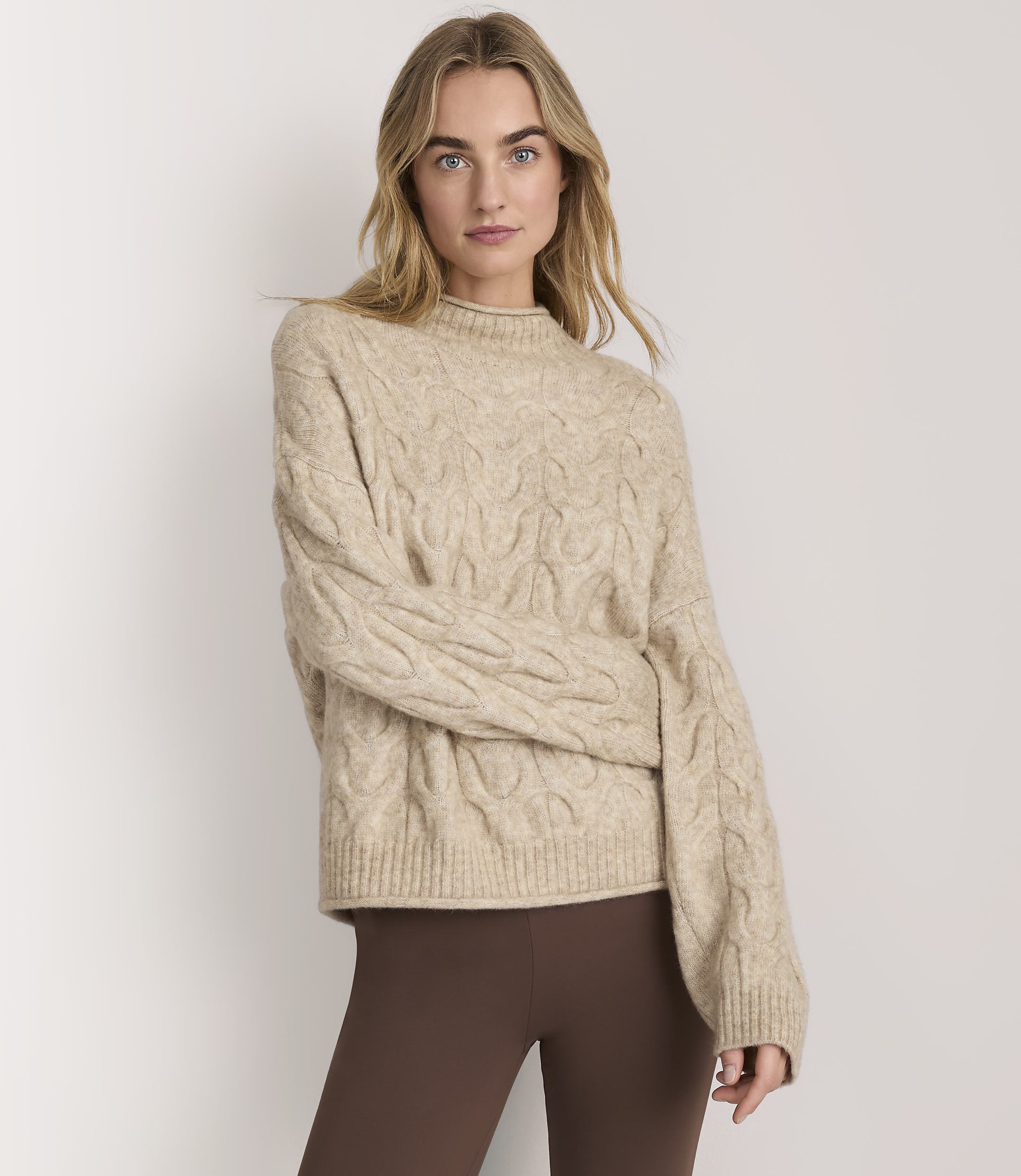 A woman wearing a beige cable-knit sweater and brown leggings stands against a plain white background.