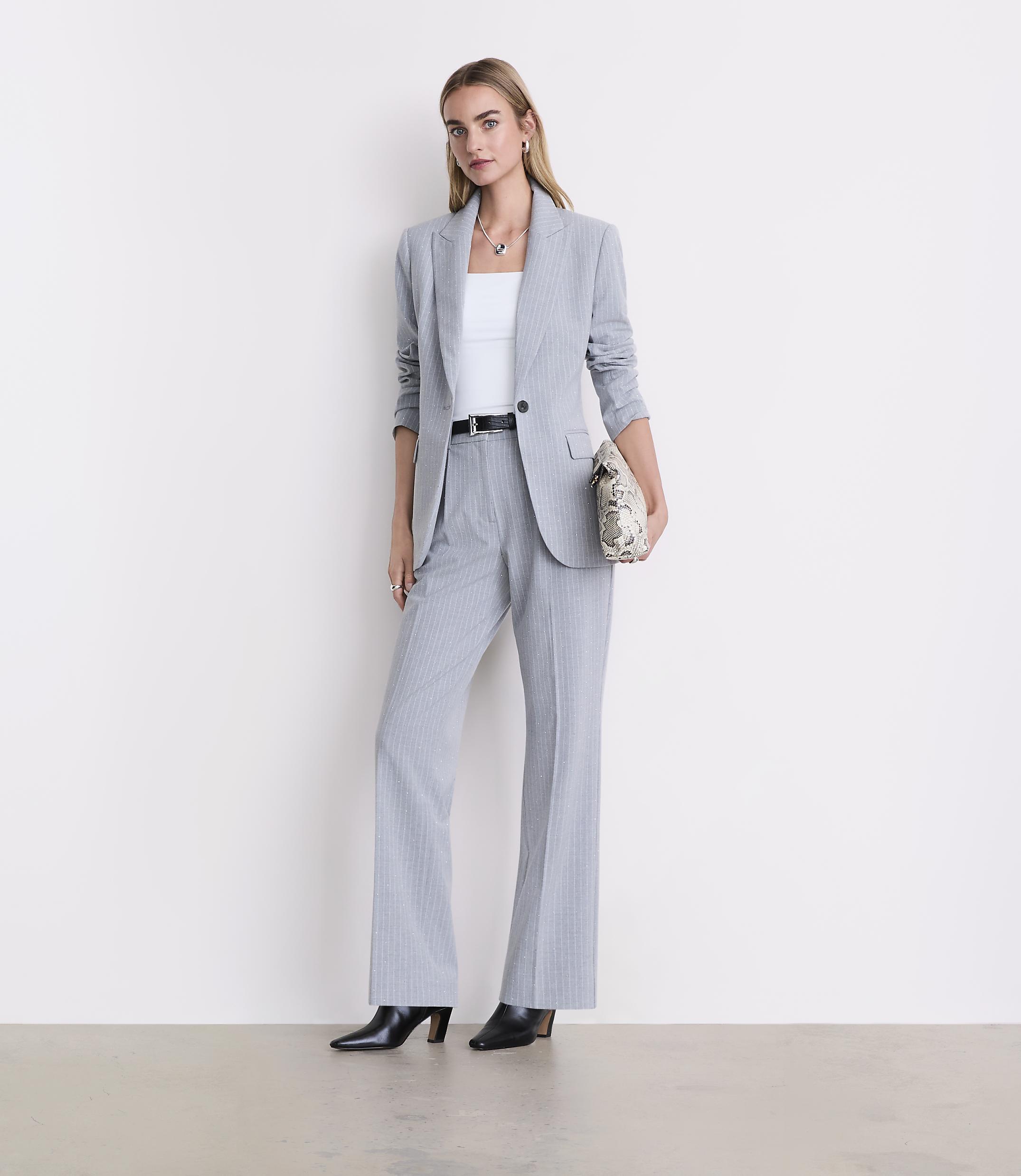 A woman wearing a light grey pinstripe suit and black heels stands against a plain white background.
