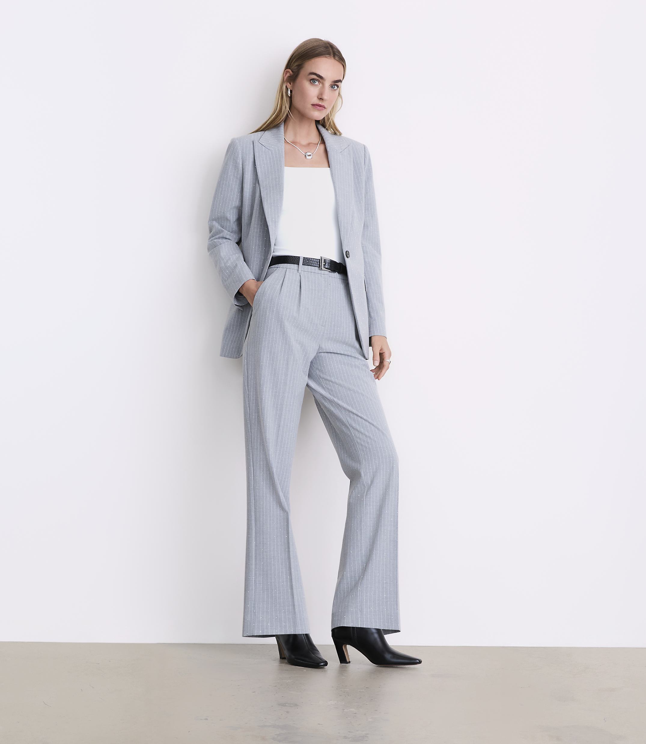 A woman wearing a light blue pinstripe suit and black heels stands against a plain white wall.