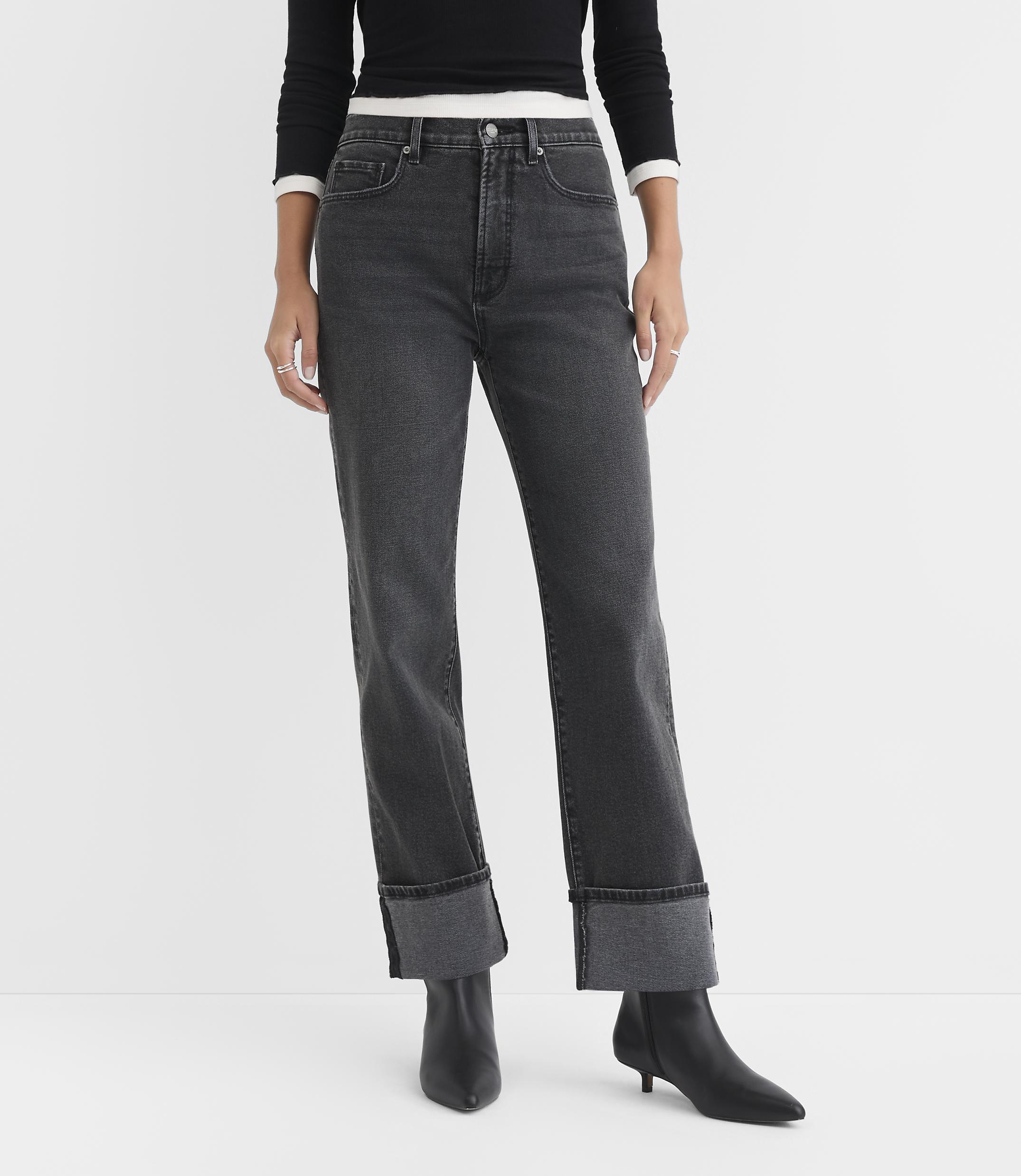 A person stands in the foreground wearing cuffed dark wash jeans and black boots against a plain white background.