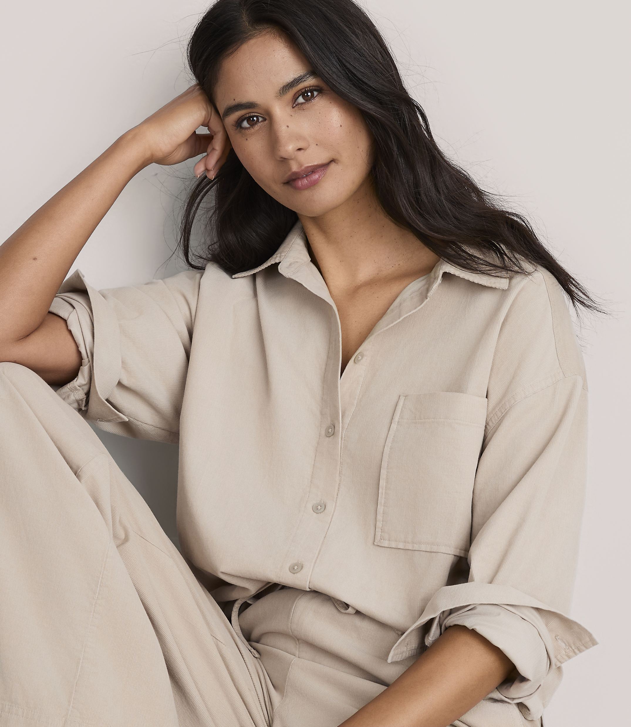 A woman with long dark hair wearing a beige button-down shirt and matching pants poses against a plain, light-colored background.